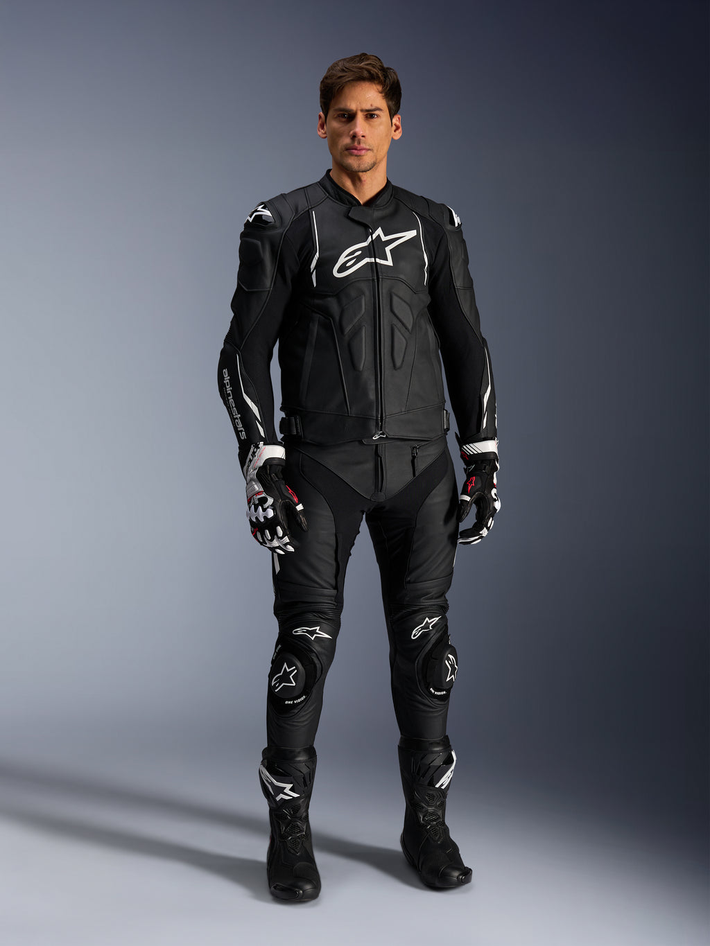 Männliches Model trägt die Alpinestars Dusk Lederjacke in Schwarz-Weiß, mit markanten weißen Logos auf der Brust und den Schultern, kombiniert mit einer passenden schwarzen Leder-Motorradhose, schwarz-weißen Handschuhen und schwarzen Stiefeln, komplettes Sport-Racing-Outfit für den Straßeneinsatz.