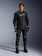 Männliches Model trägt die Alpinestars Dusk Lederjacke in Schwarz-Weiß, mit markanten weißen Logos auf der Brust und den Schultern, kombiniert mit einer passenden schwarzen Leder-Motorradhose, schwarz-weißen Handschuhen und schwarzen Stiefeln, komplettes Sport-Racing-Outfit für den Straßeneinsatz.