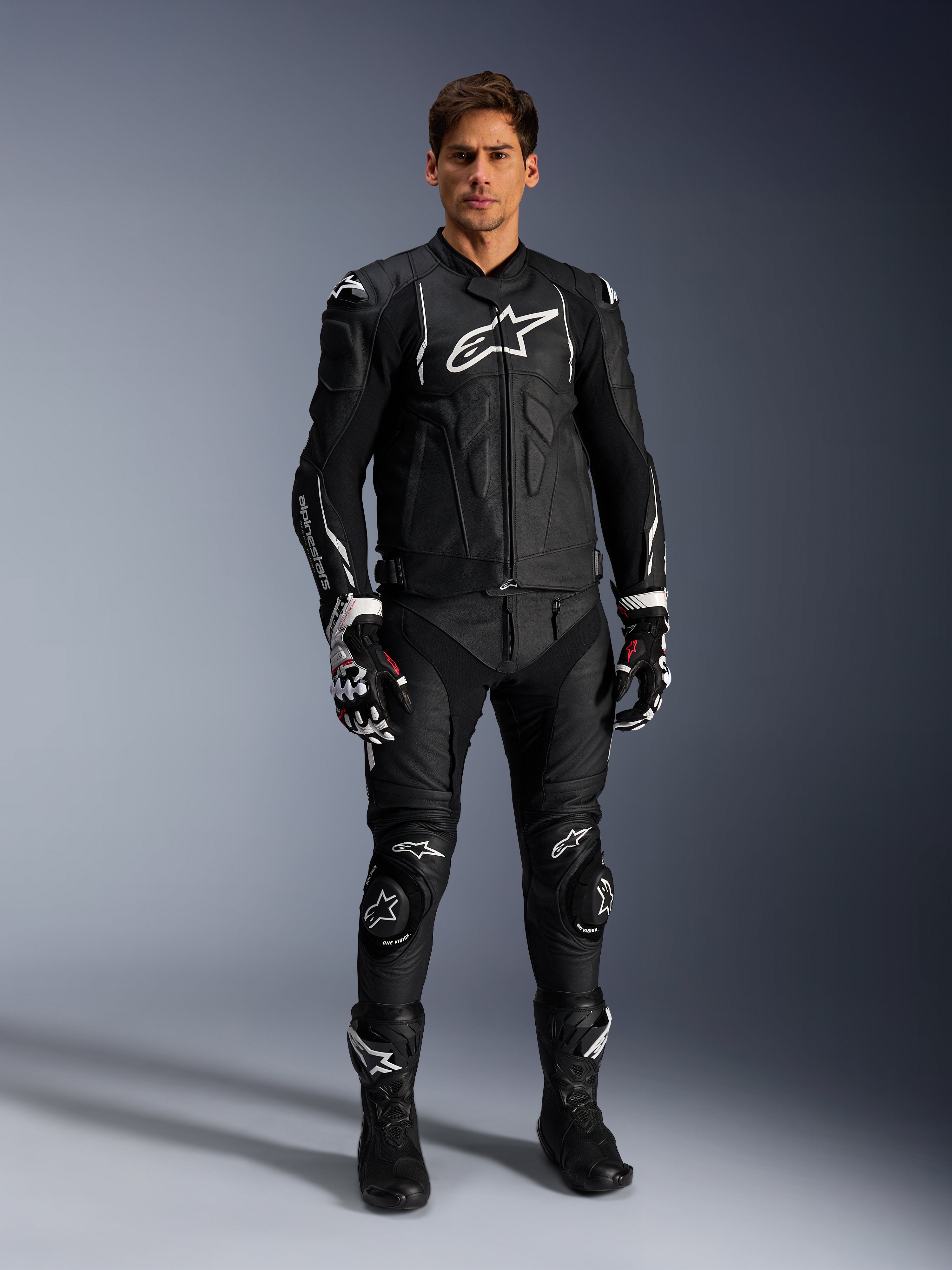 Männliches Model trägt die Alpinestars Dusk Lederjacke in Schwarz-Weiß, mit markanten weißen Logos auf der Brust und den Schultern, kombiniert mit einer passenden schwarzen Leder-Motorradhose, schwarz-weißen Handschuhen und schwarzen Stiefeln, komplettes Sport-Racing-Outfit für den Straßeneinsatz.