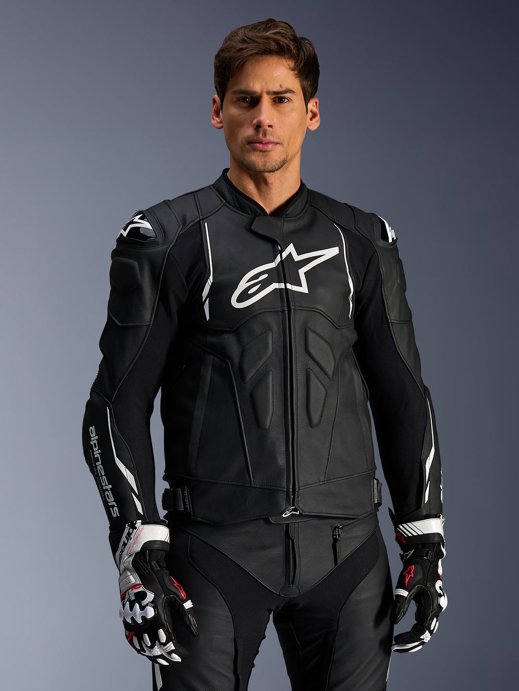 Alpinestars Dusk Lederjacke, Schwarz und Weiß, Motorrad-Lederjacke mit gepolsterten Schutzbereichen und weißen Logos, getragen mit schwarz-weißen Rennhandschuhen mit roten Akzenten, professionelle Sport-Fahrausrüstung