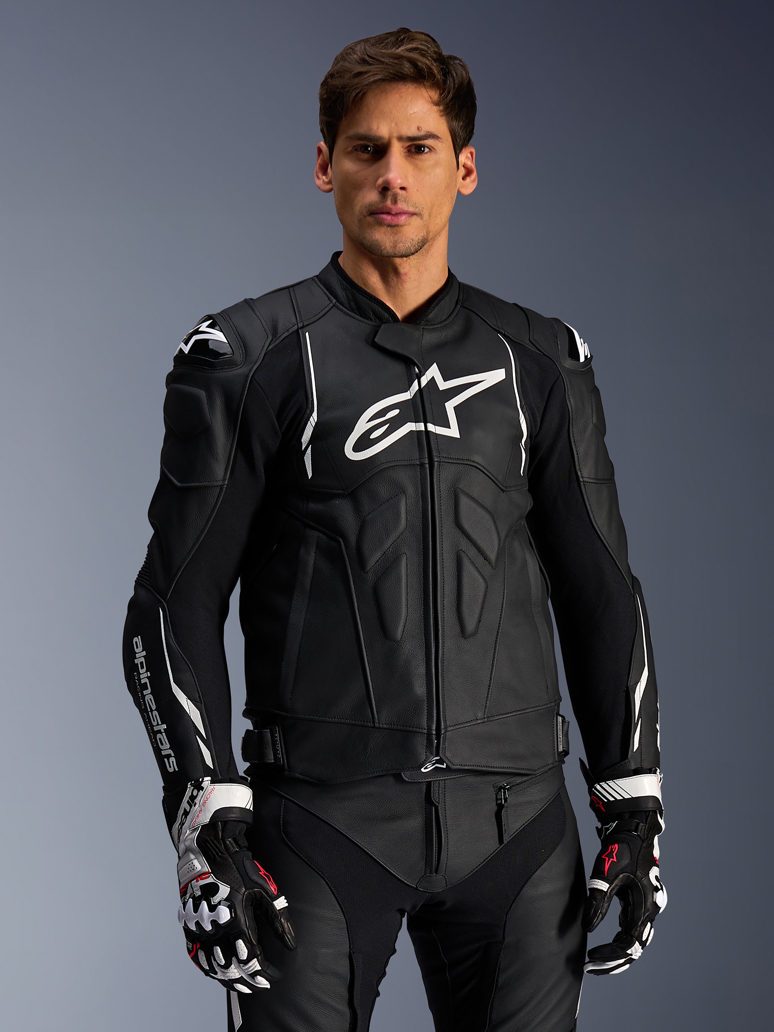 Alpinestars Dusk Lederjacke, Schwarz und Weiß, Motorrad-Lederjacke mit gepolsterten Schutzbereichen und weißen Logos, getragen mit schwarz-weißen Rennhandschuhen mit roten Akzenten, professionelle Sport-Fahrausrüstung