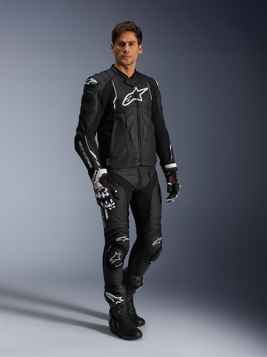 Männlicher Fahrer in der Alpinestars Dusk Lederjacke in Schwarz und Weiß, Premium-Lederjacke mit großem weißem Branding auf der Brust, kombiniert mit passender Leder-Rennhose, Handschuhen und Stiefeln, komplette Performance-Sport-Fahrausrüstung.