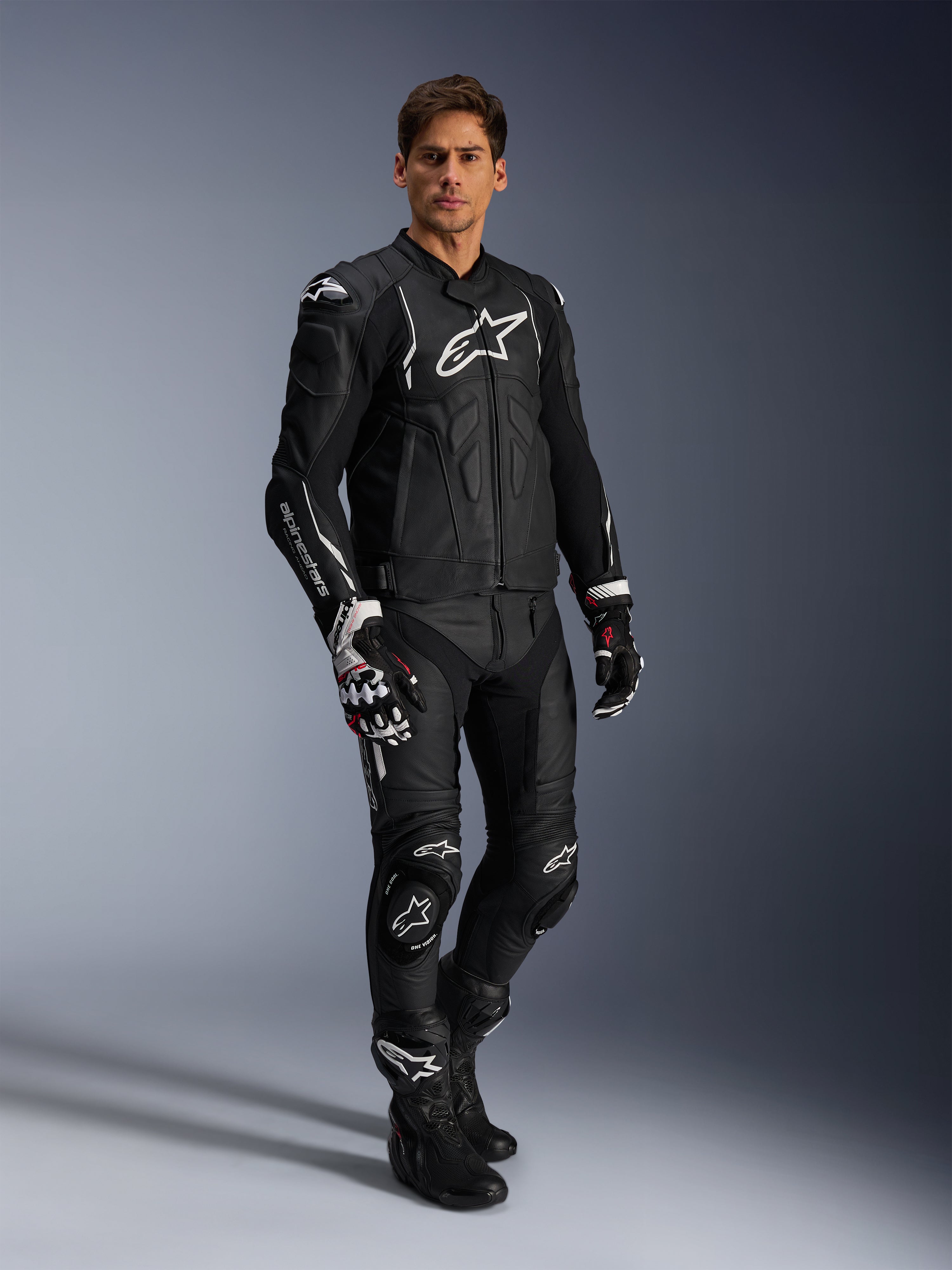 Männlicher Fahrer in der Alpinestars Dusk Lederjacke in Schwarz und Weiß, Premium-Lederjacke mit großem weißem Branding auf der Brust, kombiniert mit passender Leder-Rennhose, Handschuhen und Stiefeln, komplette Performance-Sport-Fahrausrüstung.