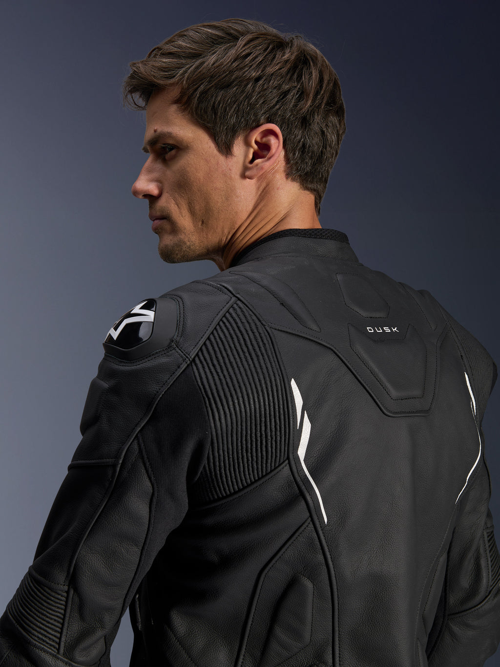 Alpinestars Dusk Lederjacke, Schwarz mit weißen Akzenten, Rückansicht am Model, mit DFS-Schulterprotektoren mit Alpinestars-Logo, Akkordeon-Leder-Stretcheinsätzen für mehr Bewegungsfreiheit und „DUSK“-Branding am oberen Rücken, Premium-Rindsleder-Motorradjacke für Straßen- und Sportfahrten