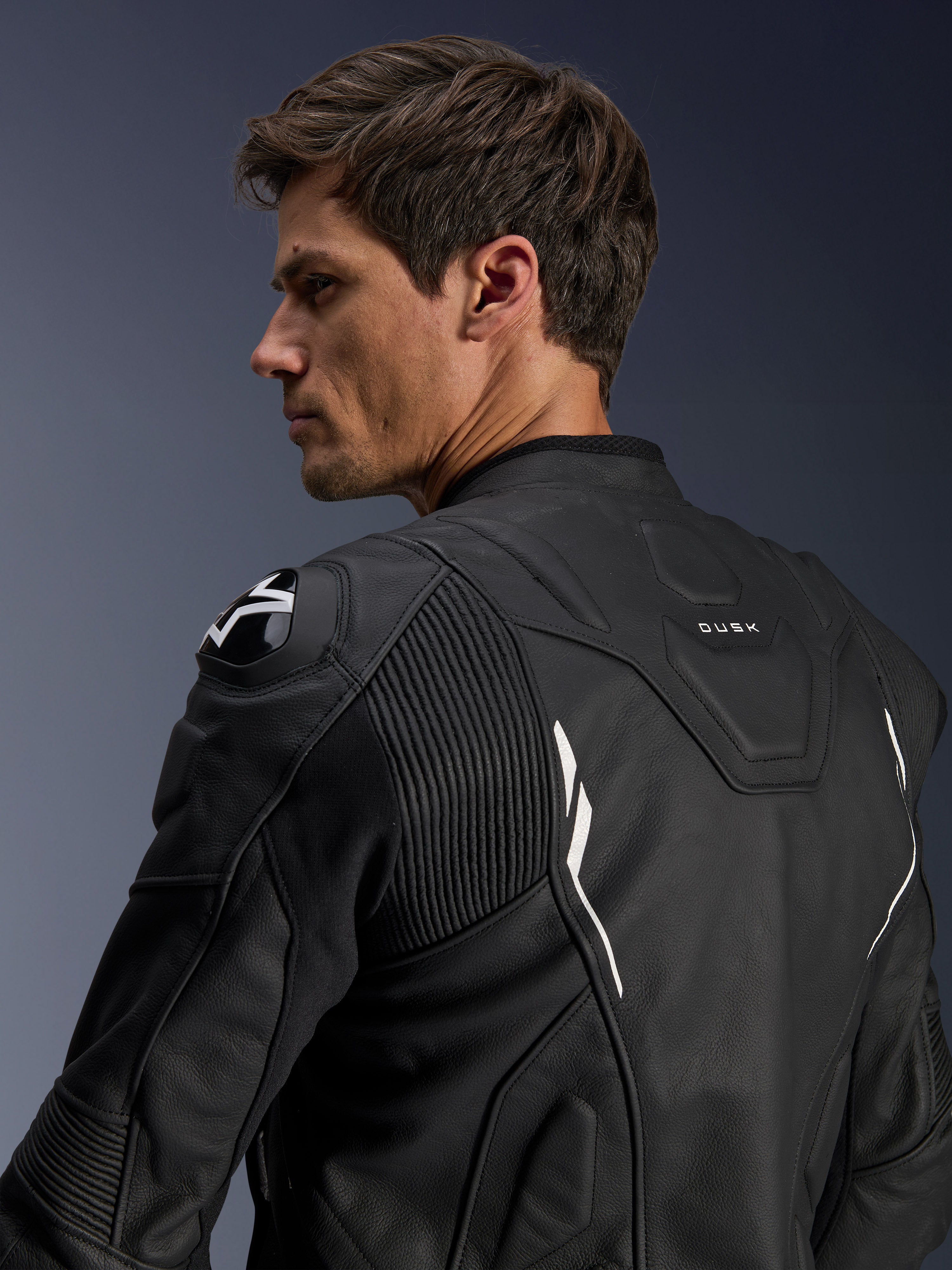 Alpinestars Dusk Lederjacke, Schwarz mit weißen Akzenten, Rückansicht am Model, mit DFS-Schulterprotektoren mit Alpinestars-Logo, Akkordeon-Leder-Stretcheinsätzen für mehr Bewegungsfreiheit und „DUSK“-Branding am oberen Rücken, Premium-Rindsleder-Motorradjacke für Straßen- und Sportfahrten