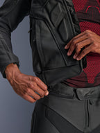 Alpinestars Dusk Lederjacke, Schwarz, Nahaufnahme des Taillen-Verbindungsreißverschlusses zur Befestigung an einer Leder-Motorradhose, getragen mit einem rot-schwarz gemusterten Baselayer, strapazierfähige Lederkonstruktion für den Straßeneinsatz