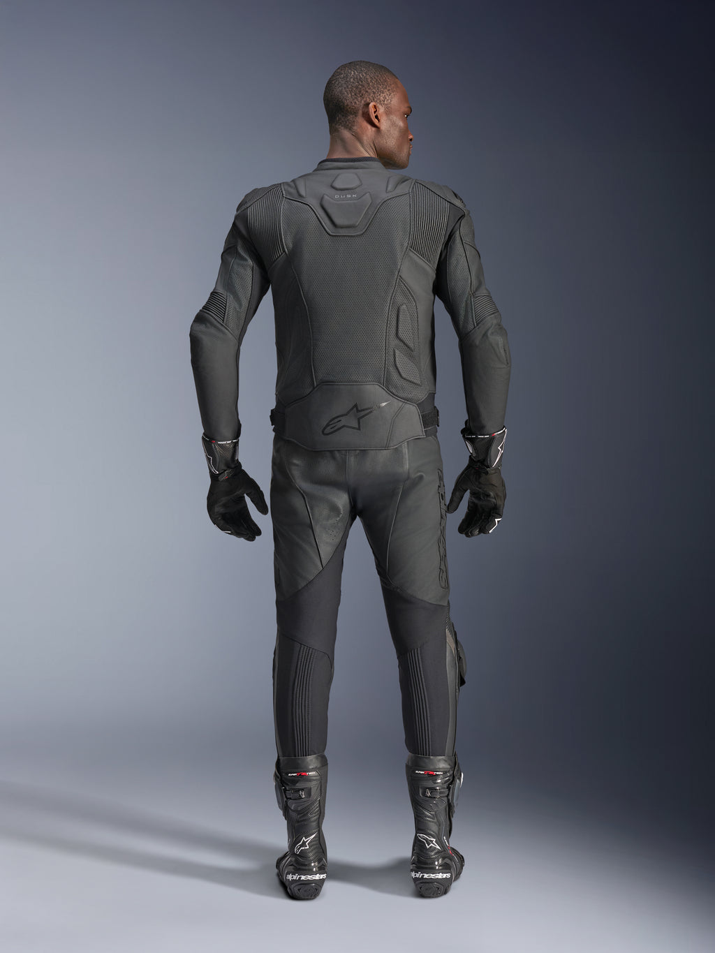 Männliches Model in der Rückansicht mit der Alpinestars Dusk Airflow Lederjacke in Schwarz, kombiniert mit passender schwarzer Leder-Rennhose, Handschuhen und Stiefeln, mit großflächigen Perforationen zur Belüftung und dem Alpinestars-Logo auf dem unteren Rücken.
