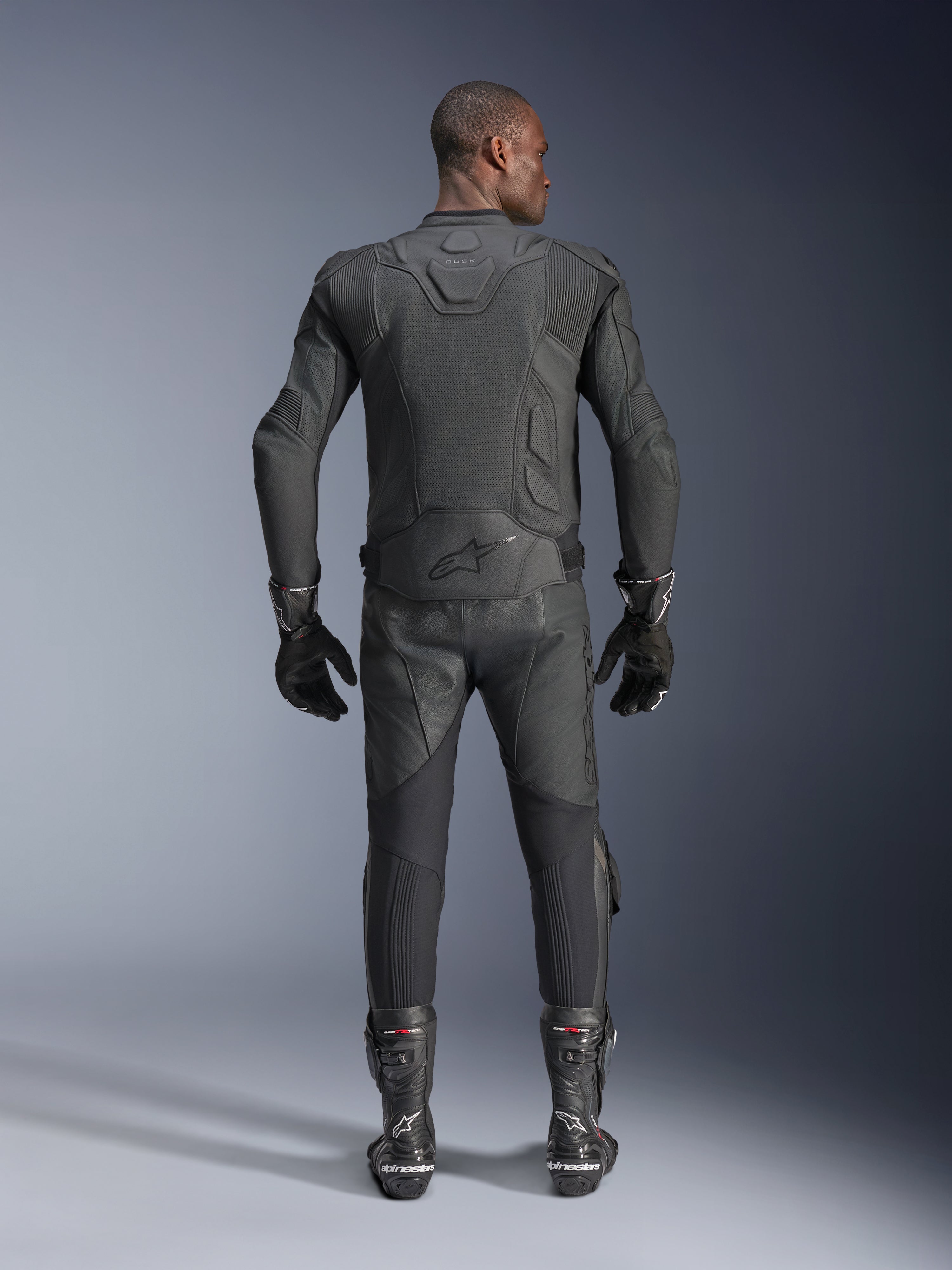 Männliches Model in der Rückansicht mit der Alpinestars Dusk Airflow Lederjacke in Schwarz, kombiniert mit passender schwarzer Leder-Rennhose, Handschuhen und Stiefeln, mit großflächigen Perforationen zur Belüftung und dem Alpinestars-Logo auf dem unteren Rücken.
