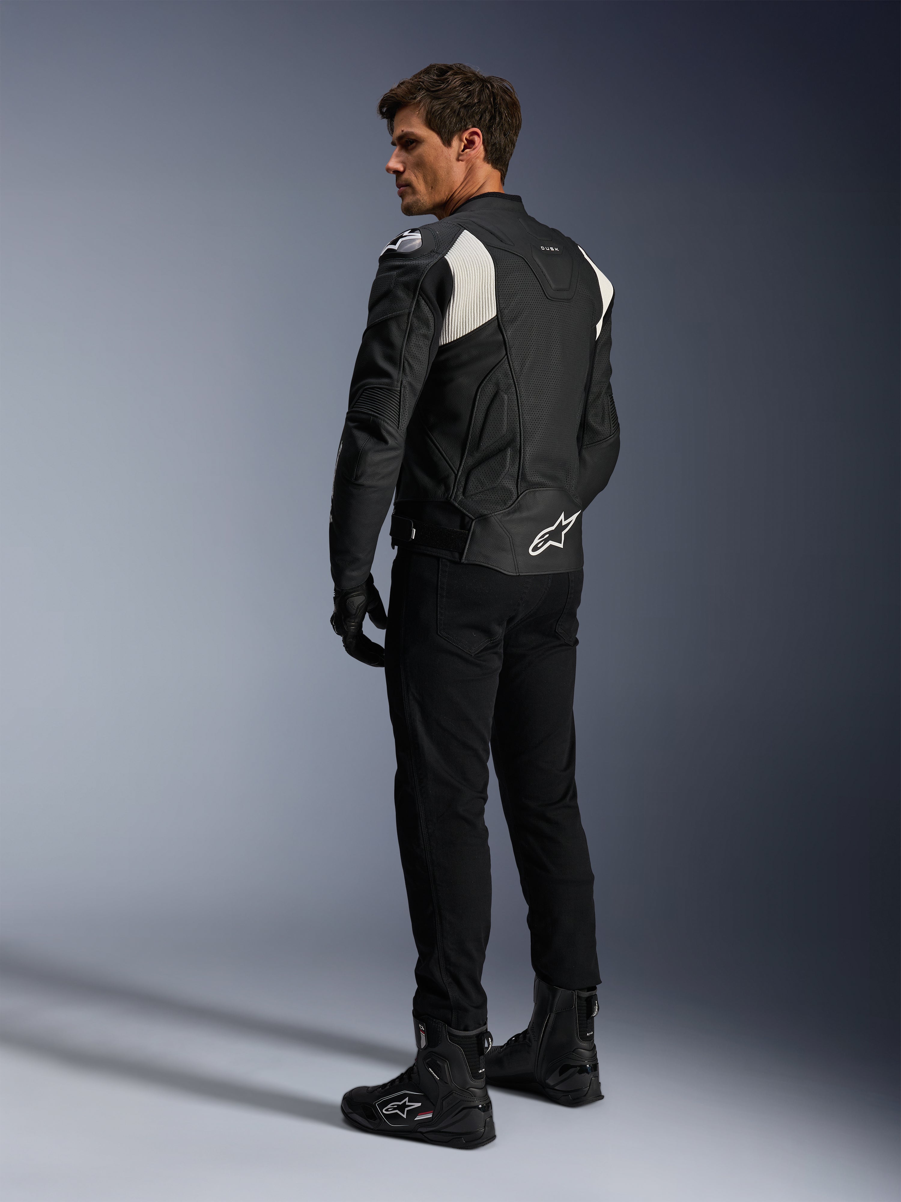 Alpinestars Dusk Airflow Lederjacke, Schwarz und Weiß, Rückansicht eines Models in der perforierten Lederjacke mit passender schwarzer Hose und Stiefeln, mit weißen Akkordeon-Stretcheinsätzen und markantem Logo am unteren Rücken, Sport-Motorradbekleidung