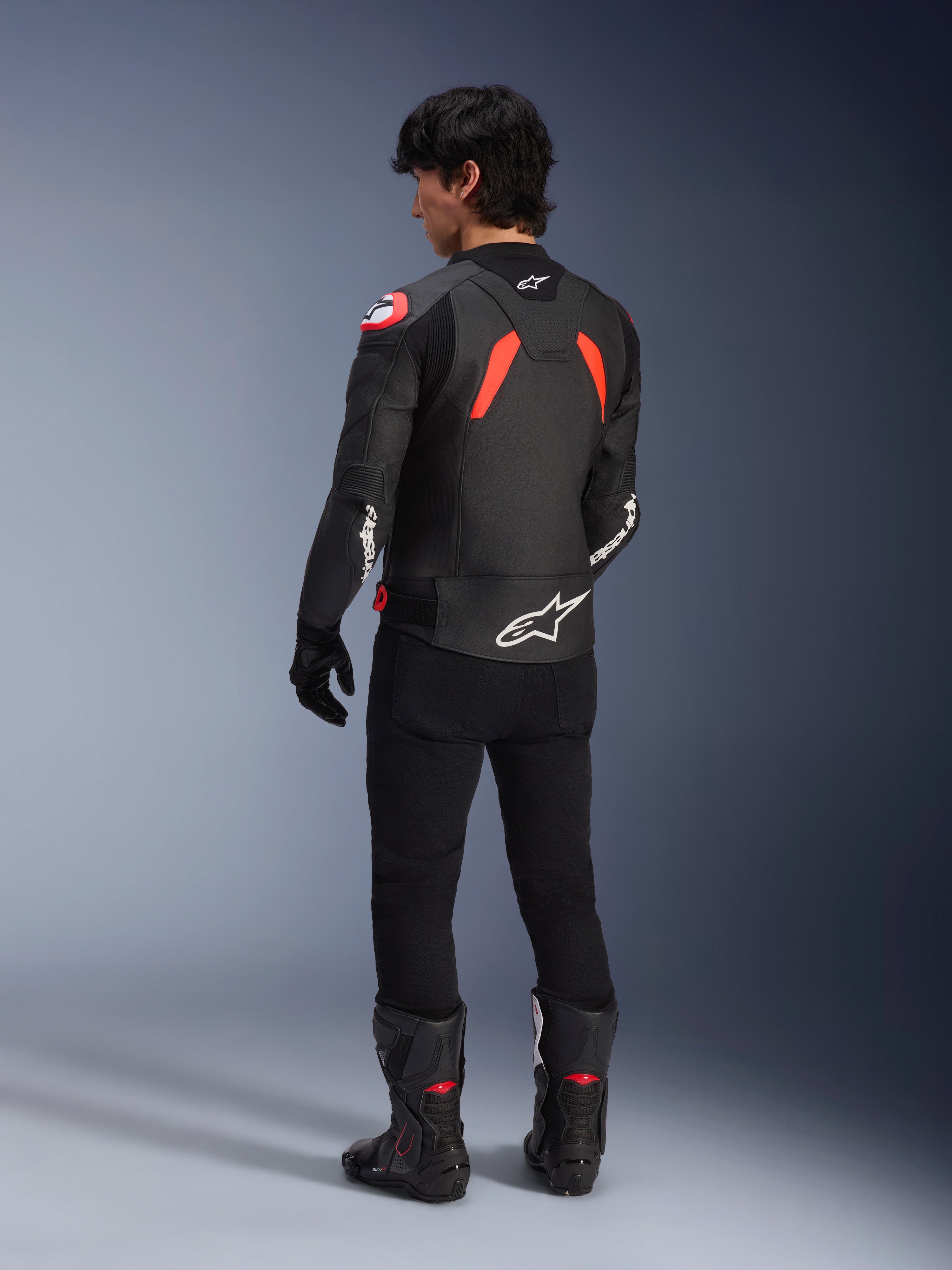 Rear view of a model wearing the Alpinestars GP Plus R V4 Airflow Leather Jacket, Black with Red Fluo and White accents, worn with black pants and black motorcycle boots, featuring prominent white logos on the back and sleeves, sport-style leather construction
