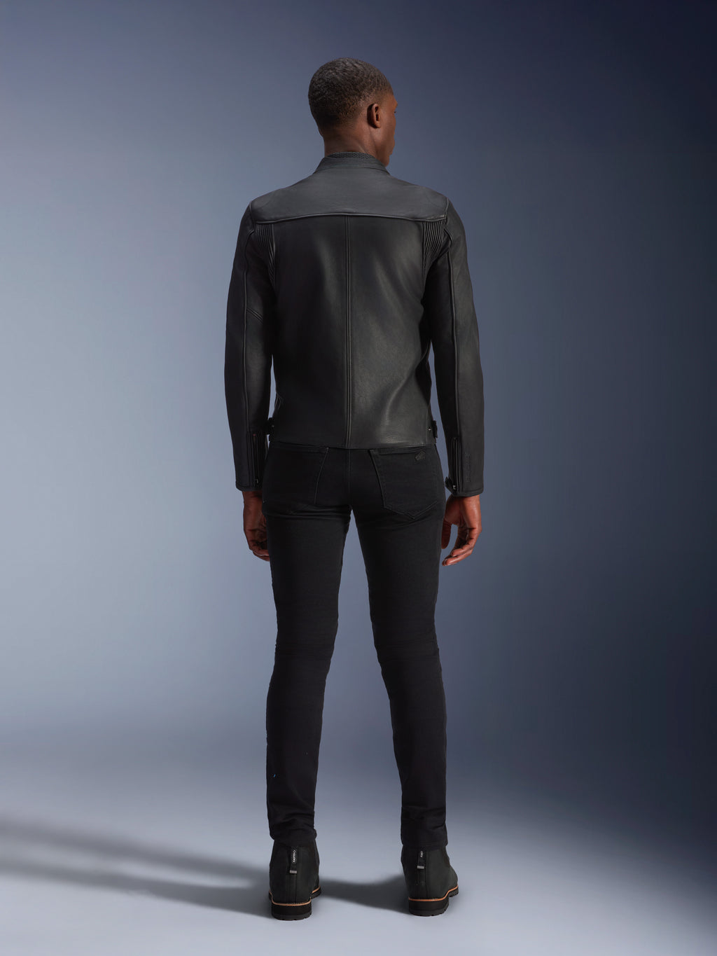 Alpinestars Blacktrack Leather Jacket, rear view, all black premium bovine leather, featuring accordion panels on shoulder blades for mobility, worn with black slim-fit pants and black boots, urban contemporary motorcycle gear.