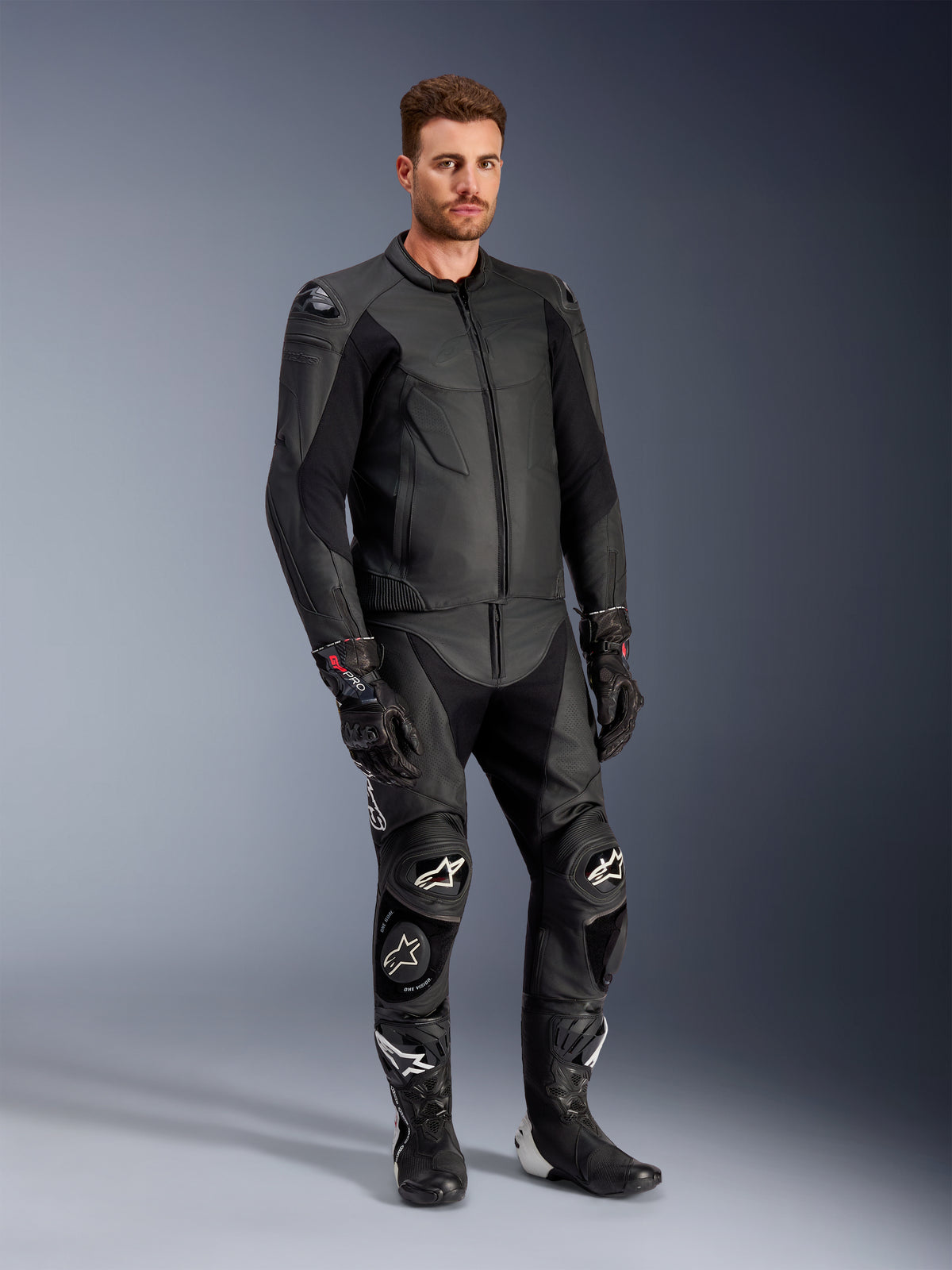Männliches Modell in der Alpinestars Caliber Lederjacke in Schwarz, kombiniert mit passender schwarzer Leder-Motorradhose, Rennhandschuhen und Stiefeln, präsentiert ein komplettes Sport-Road-Outfit.