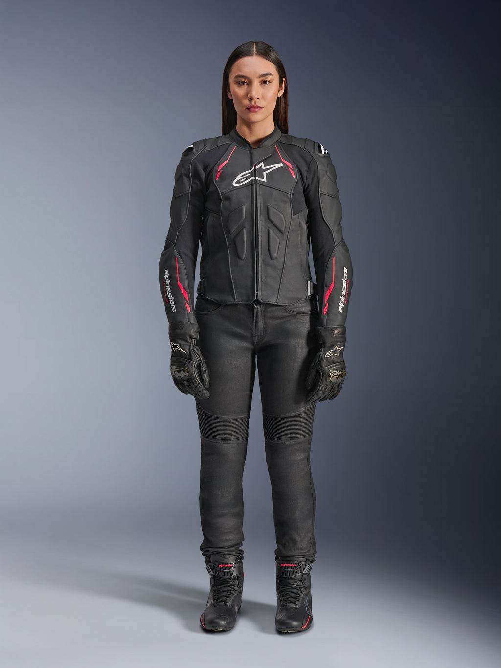 A woman wearing the Alpinestars Stella Dusk Leather Jacket in Black Diva Pink White, featuring a white chest logo and pink accents on the shoulders and sleeves, paired with black motorcycle pants, gloves, and boots.