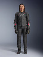 A woman wearing the Alpinestars Stella Dusk Leather Jacket in Black Diva Pink White, featuring a white chest logo and pink accents on the shoulders and sleeves, paired with black motorcycle pants, gloves, and boots.