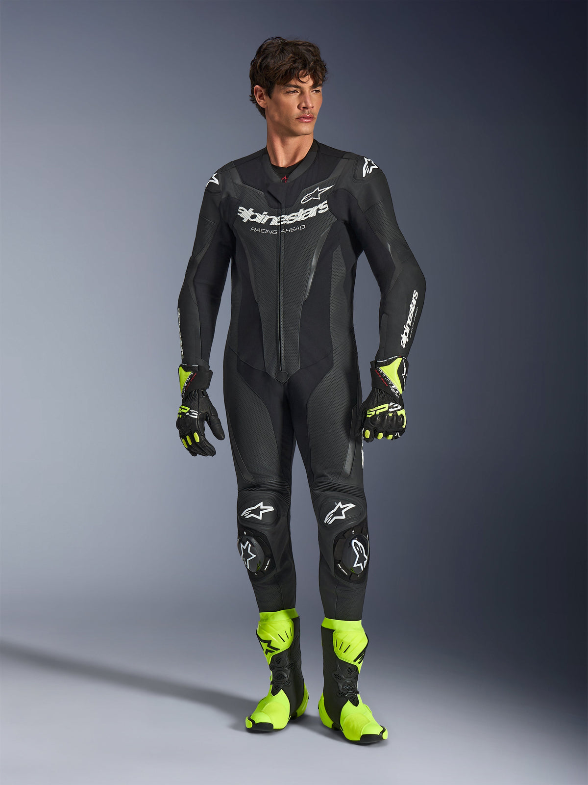 Alpinestars GP Force V2 Lederkombi, einteiliger schwarzer Motorradrennanzug, getragen mit neongelben Stiefeln und Handschuhen, mit weißen Logos und perforiertem Leder, schlankes Design für Trackdays