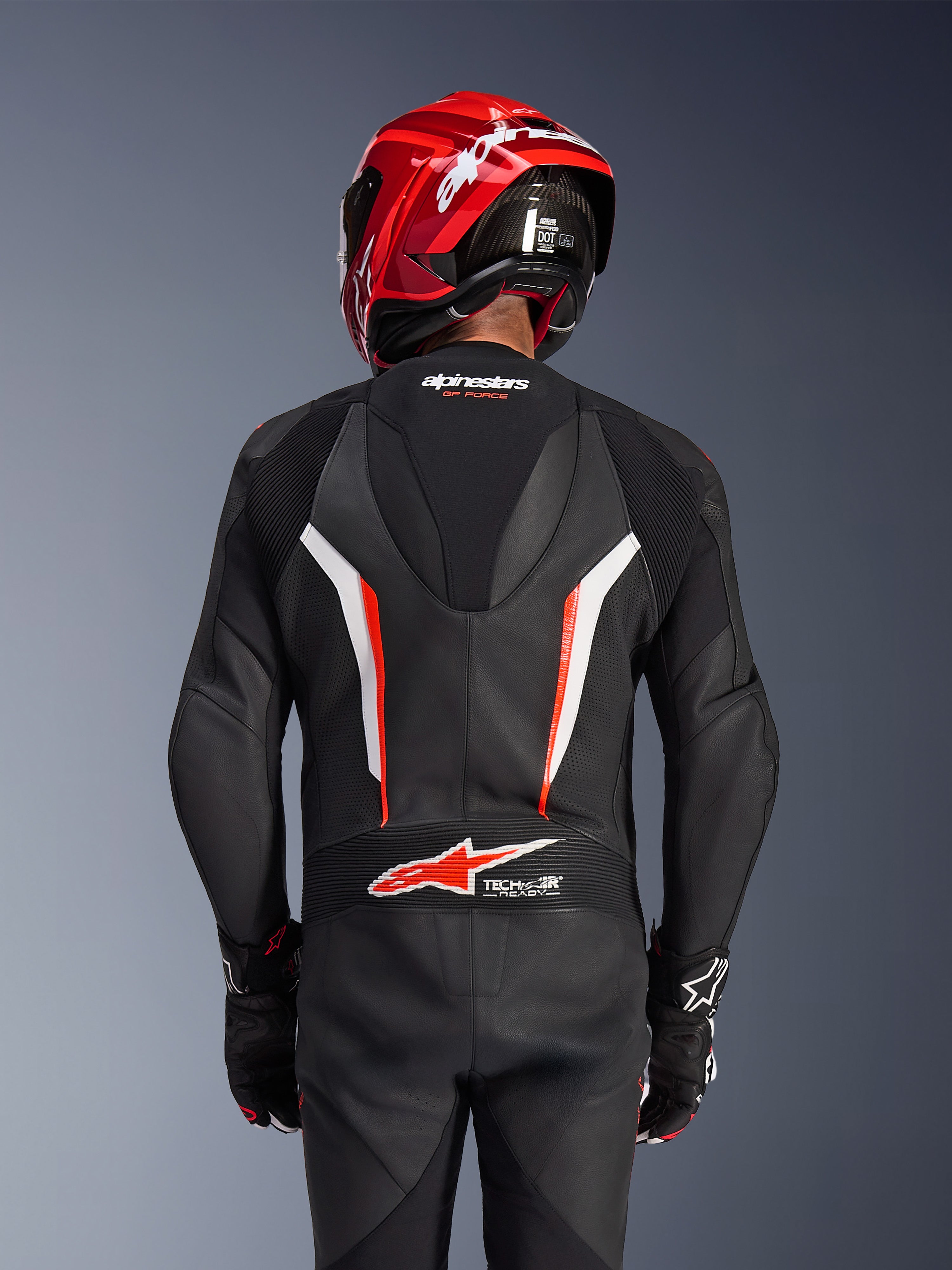 Alpinestars GP Force V2 Leather Suit 1-Piece, Black White and Red Fluo, back view of rider wearing red racing helmet and gloves, featuring perforated leather panels, aerodynamic speed hump with Alpinestars branding, and Tech-Air Ready logo on the lower back, professional motorcycle racing gear.