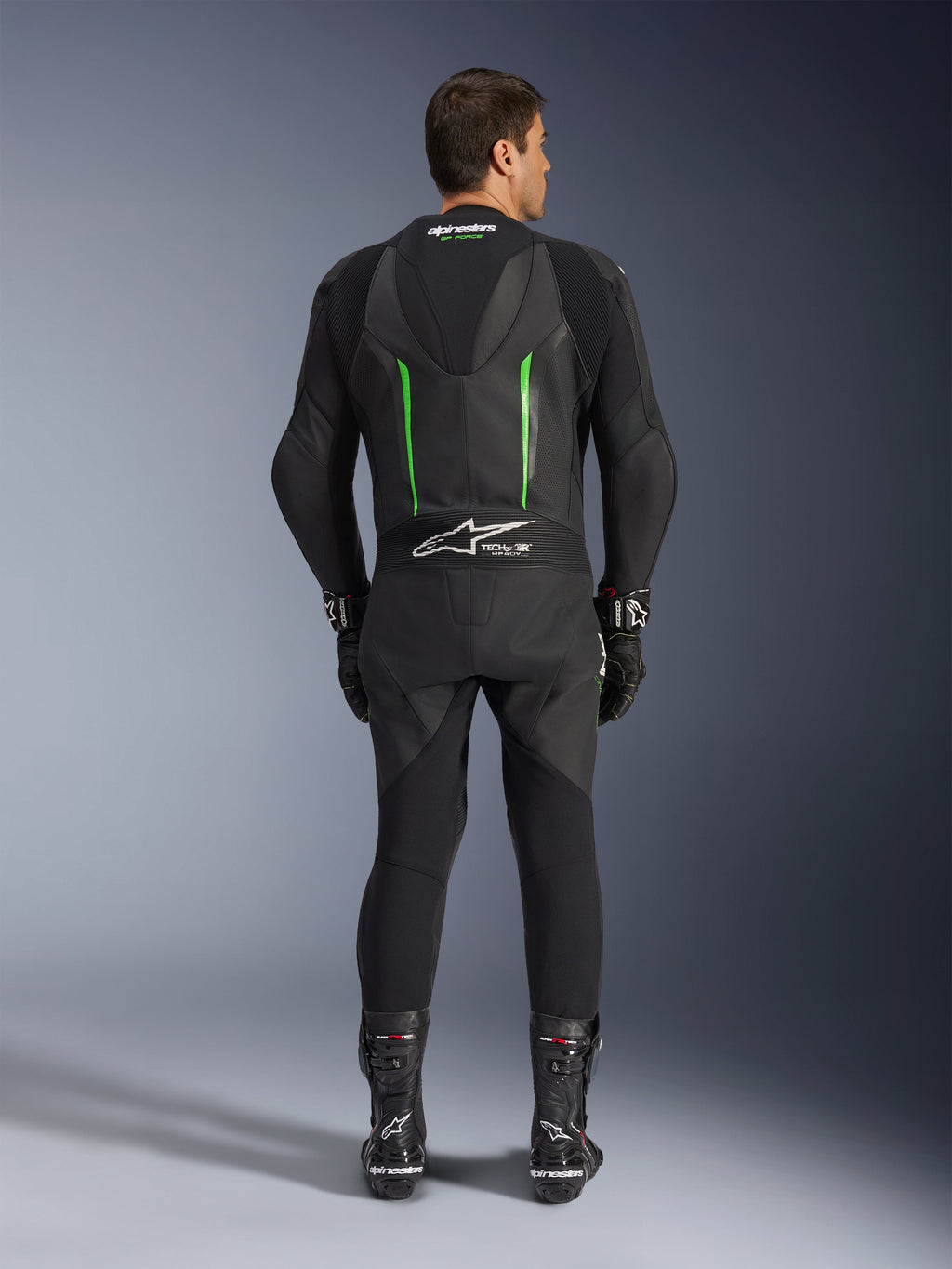 Rear view of the Alpinestars GP Force V2 1-Piece Leather Suit in Black and Green, featuring an aerodynamic speed hump, perforated panels, and Tech-Air Ready branding on the lower back, worn with black racing gloves and boots.