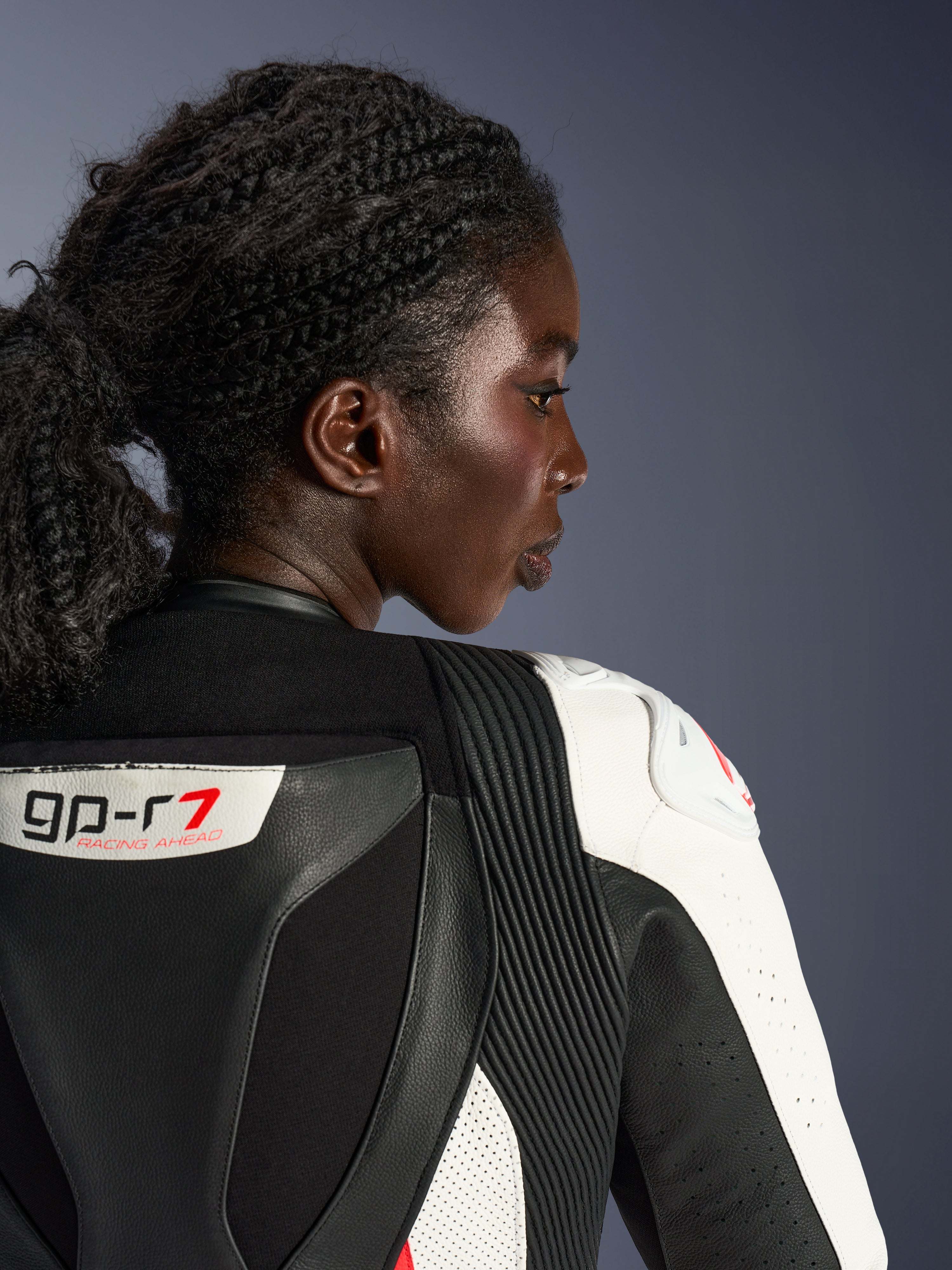 Close-up profile of a woman wearing the Alpinestars Stella GP-R7 2-Piece Leather Suit, showing the upper back and shoulder area in black and white leather, featuring the 'GP-R7' logo, accordion stretch panels, and white shoulder armor with a red logo.