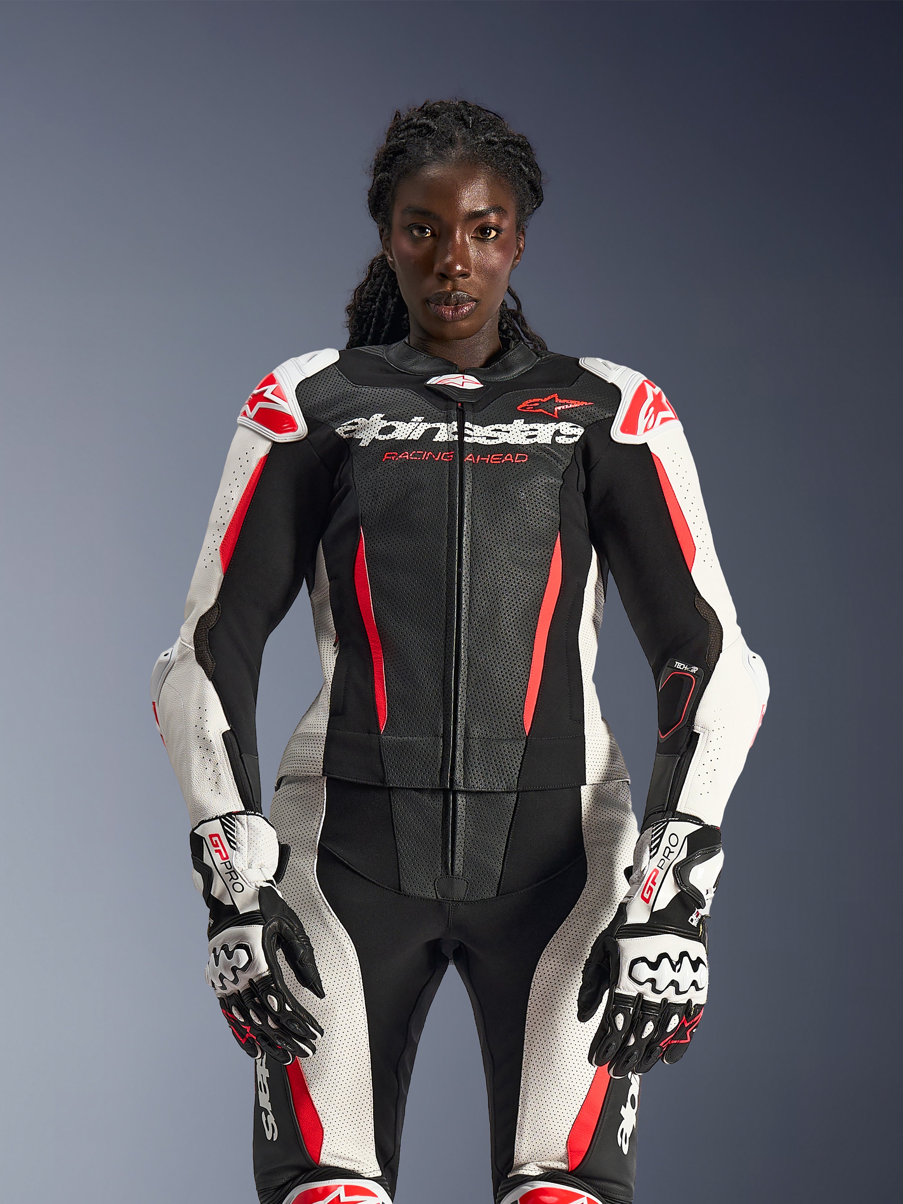 Female model wearing the Alpinestars Stella GP-R7 2-Piece Leather Suit in Black, White, and Red Fluo, featuring perforated leather panels and matching GP Pro gloves, anatomically optimized for women riders.