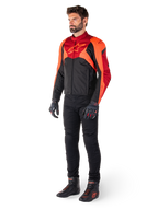 Male rider wearing the Alpinestars T-Jaws V4 Waterproof Jacket in Black Red Fluo Dark Red, paired with black motorcycle pants, gloves, and riding boots, featuring a sport-fit textile design with bold chest branding and an aggressive silhouette.