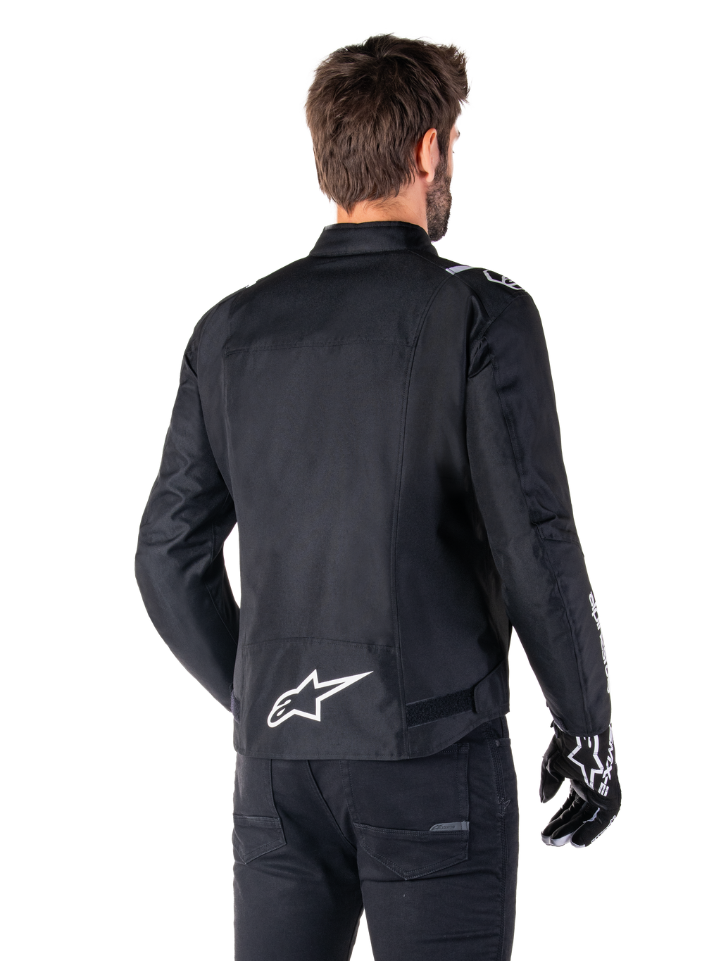 Alpinestars T-SPS V2 Wasserdichte Jacke, Schwarz und Weiß, Rückansicht eines männlichen Fahrers, der die Jacke trägt, weißes Alpinestars-Logo auf dem unteren Rücken, Textilkonstruktion im Sportschnitt, getragen mit schwarzer Motorradhose und Handschuhen