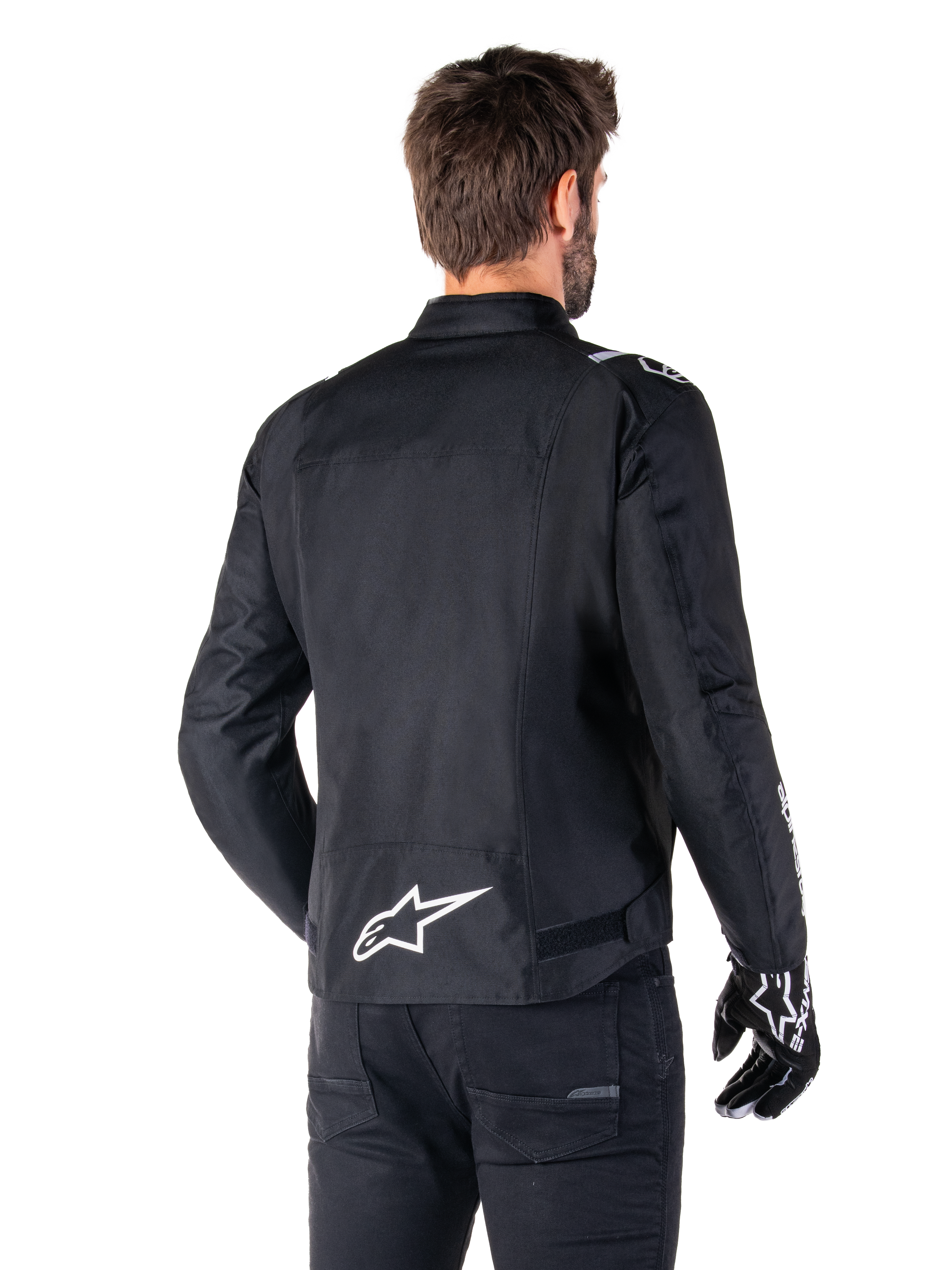 Alpinestars T-SPS V2 Wasserdichte Jacke, Schwarz und Weiß, Rückansicht eines männlichen Fahrers, der die Jacke trägt, weißes Alpinestars-Logo auf dem unteren Rücken, Textilkonstruktion im Sportschnitt, getragen mit schwarzer Motorradhose und Handschuhen