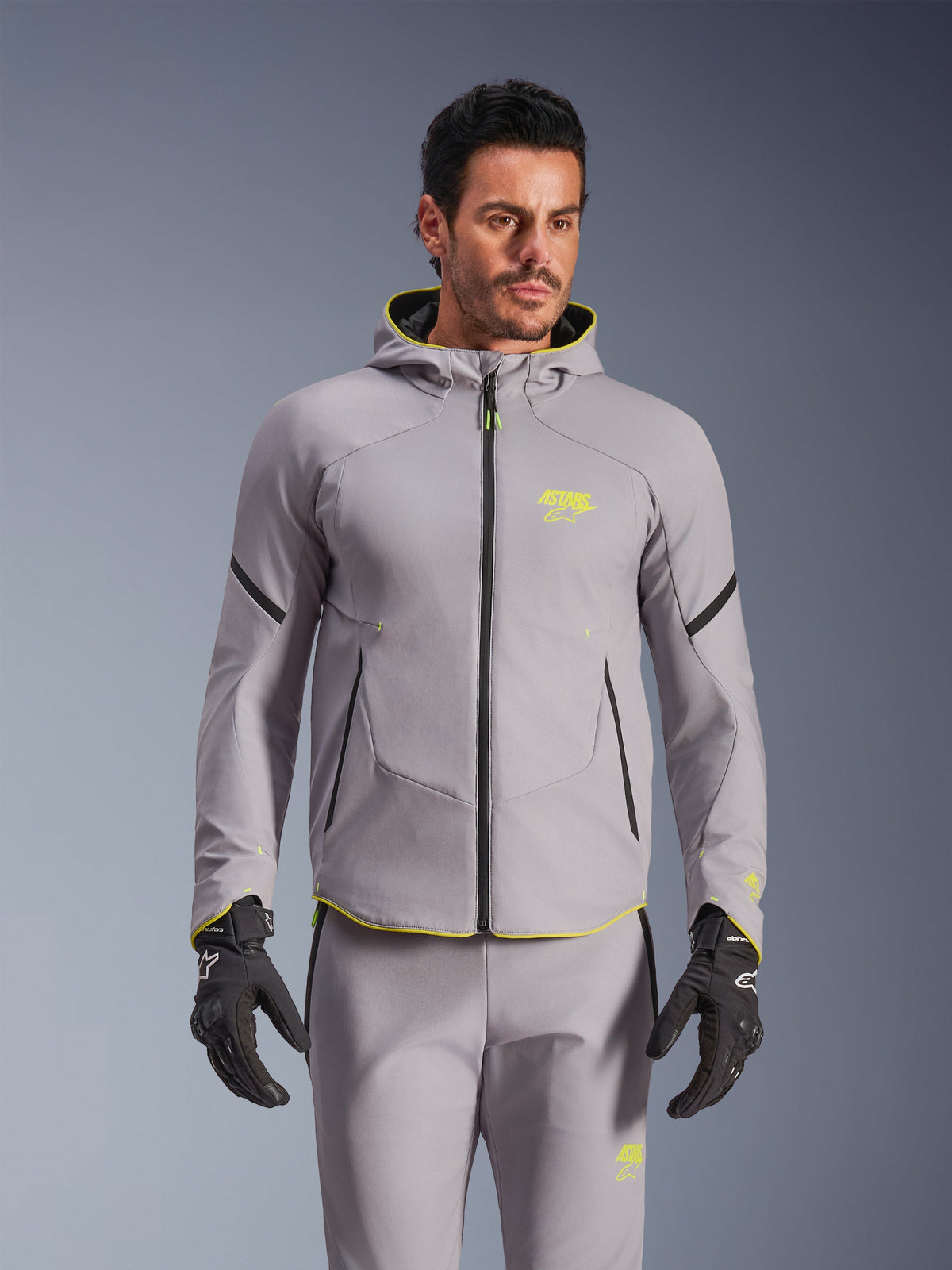 Alpinestars Aeroshell Waterproof Jacket, Mittelgrau und Gelb Fluo, Motorrad-Softshelljacke mit Kapuze und schwarzen Reißverschlüssen, getragen mit passender grauer Hose und schwarzen Handschuhen, wasserdichte Funktionsbekleidung für den Straßeneinsatz
