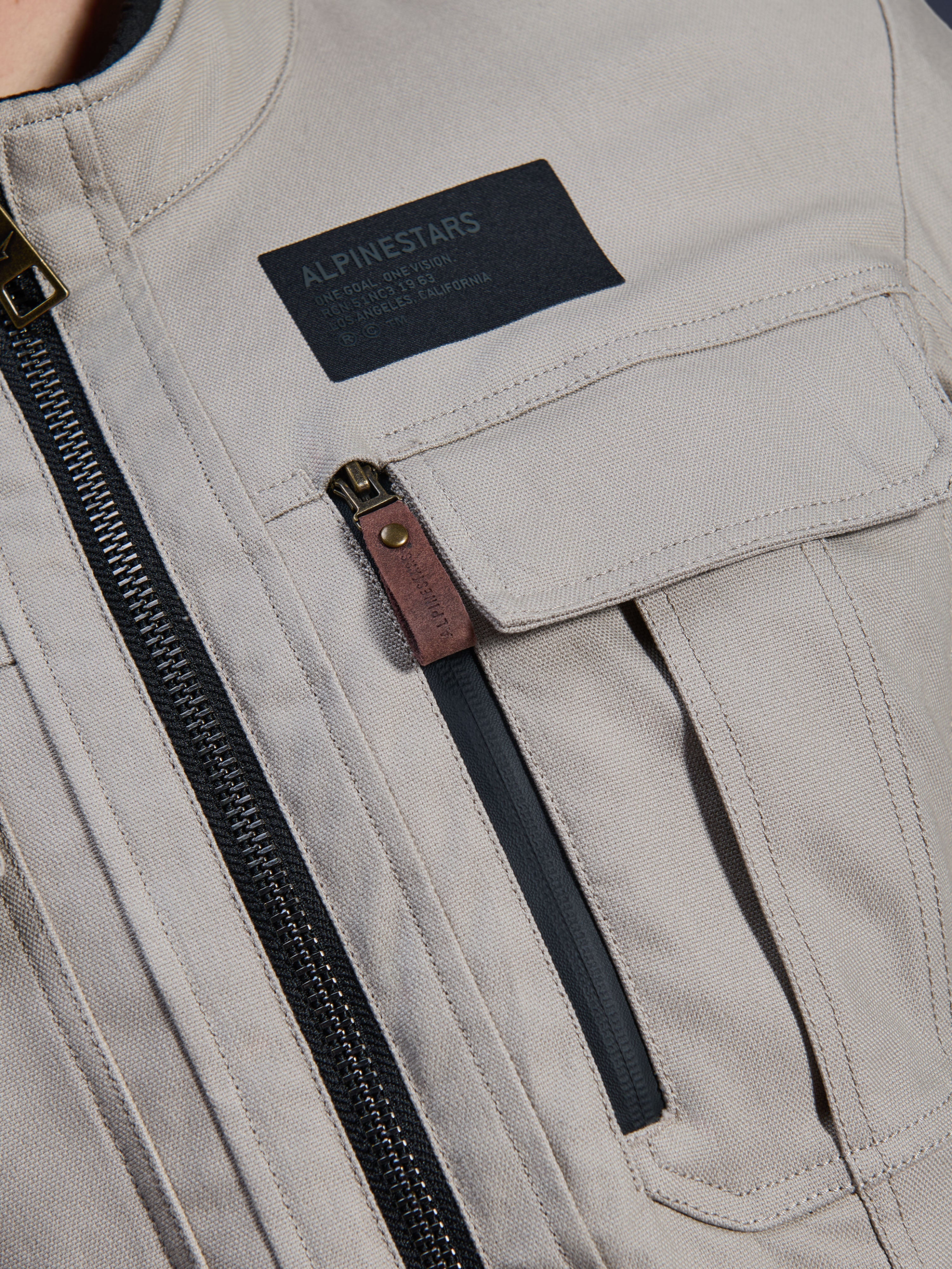 Alpinestars Borrego Drystar® Jacket, Warm Gray, close-up detail of chest area featuring a black Alpinestars branding patch, chest pocket with flap, and a vertical zipper with a brown leather pull tab, durable gray textile canvas construction.