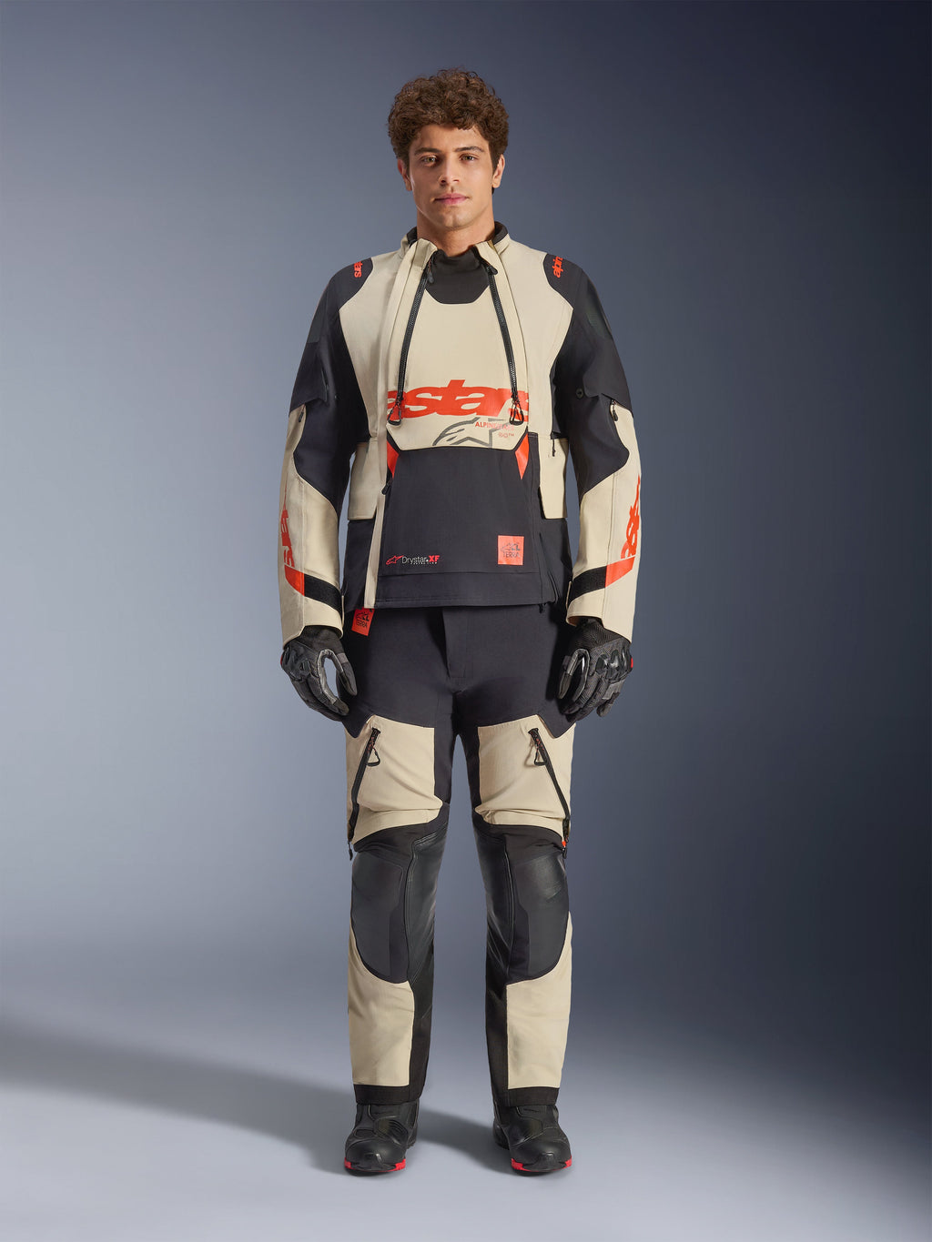 Alpinestars Halo Pro Drystar® XF Laminierte Jacke, Mastic Black, Adventure-Motorradjacke getragen von einem Model mit passender Hose und Handschuhen, beige und schwarze Textilpaneele mit markanten roten Logos, modulares Design für Fahrten in jedem Gelände
