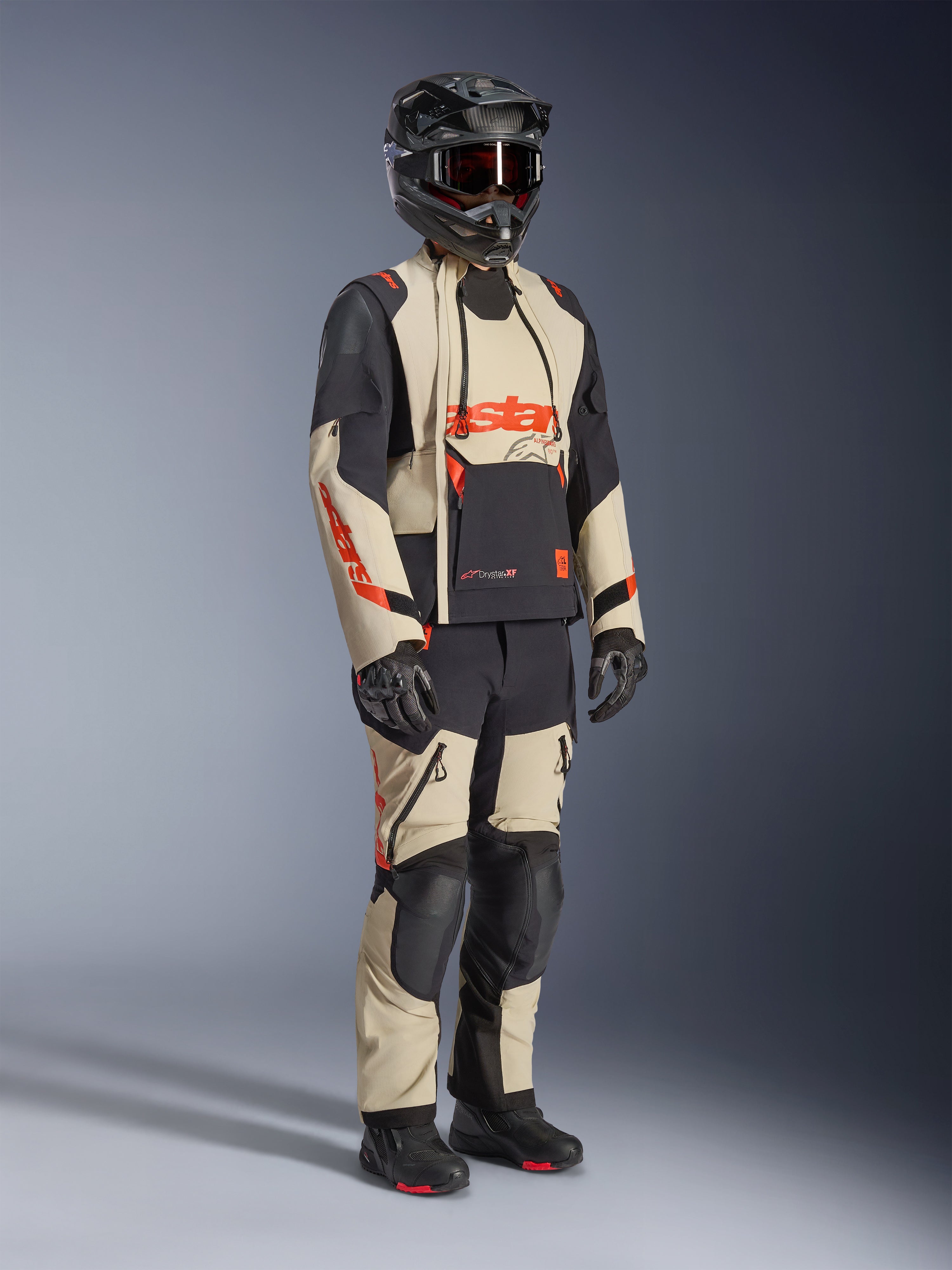 Alpinestars Halo Pro Drystar® XF Laminierte Jacke in Mastic Black, Adventure-Motorradjacke getragen mit passender Hose, Integralhelm, Handschuhen und Stiefeln, mit beigen und schwarzen Einsätzen und roten Logos, technisches Mehrjahreszeiten-Design.