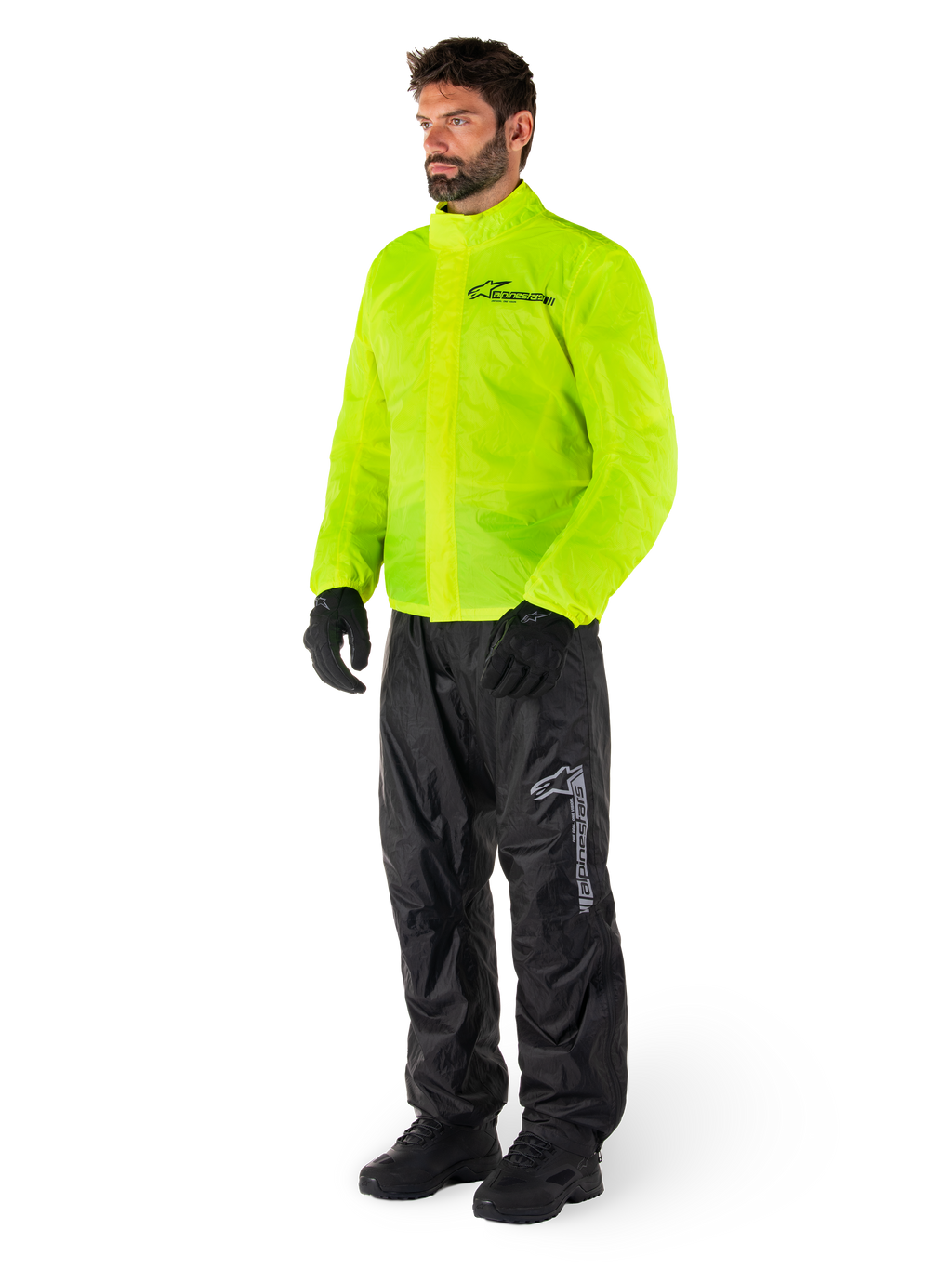 Mann trägt Alpinestars Hurricane Rain V2 Jacke in Neongelb und Hurricane Rain V2 Hose in Schwarz, hochsichtbare wasserdichte Motorrad-Regenbekleidung, mit reflektierenden Logos auf der Brust und am Oberschenkel, komplettes Outfit mit schwarzen Handschuhen und Stiefeln.