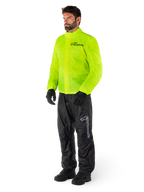 Mann trägt Alpinestars Hurricane Rain V2 Jacke in Neongelb und Hurricane Rain V2 Hose in Schwarz, hochsichtbare wasserdichte Motorrad-Regenbekleidung, mit reflektierenden Logos auf der Brust und am Oberschenkel, komplettes Outfit mit schwarzen Handschuhen und Stiefeln.