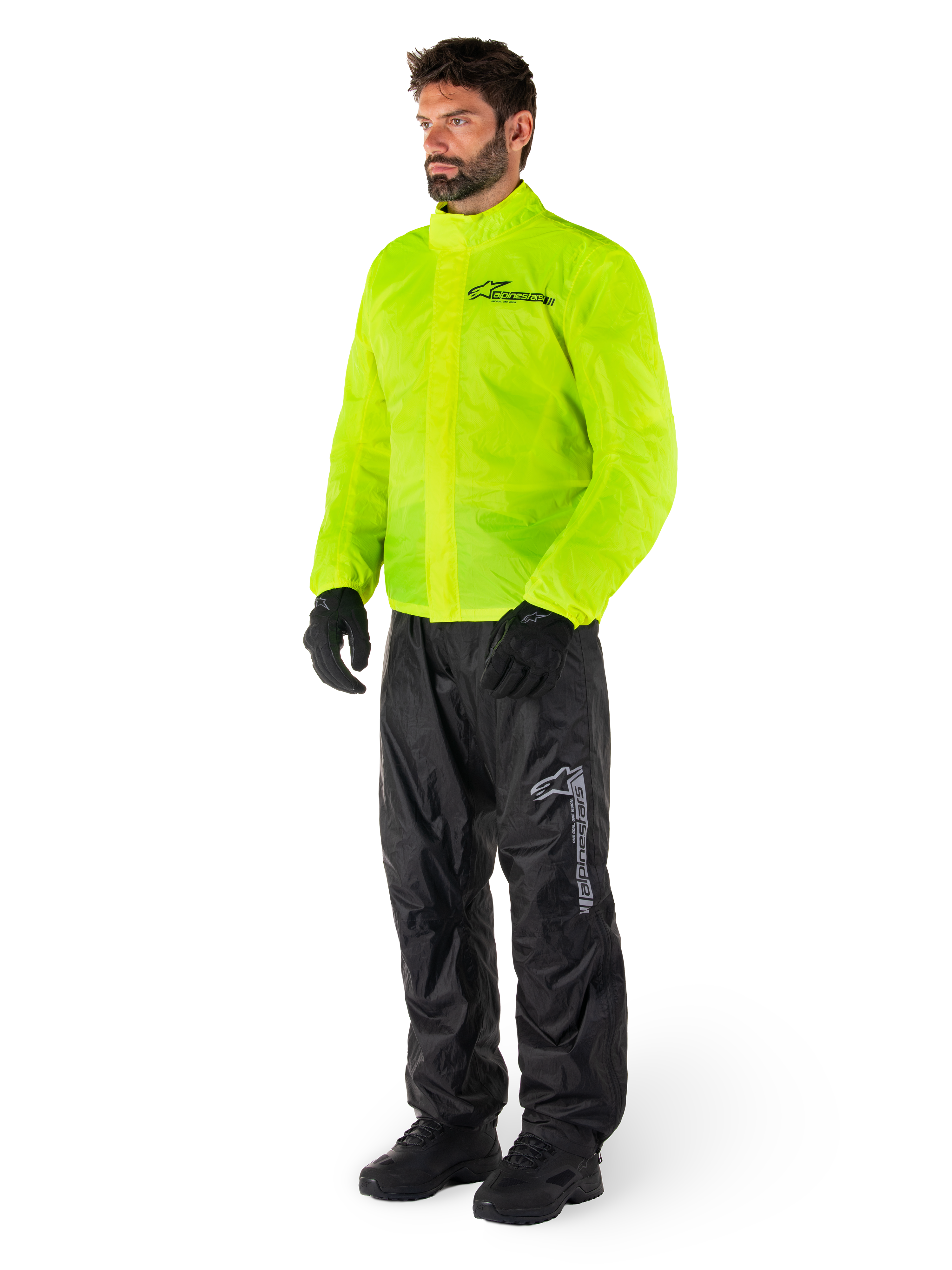 Mann trägt Alpinestars Hurricane Rain V2 Jacke in Neongelb und Hurricane Rain V2 Hose in Schwarz, hochsichtbare wasserdichte Motorrad-Regenbekleidung, mit reflektierenden Logos auf der Brust und am Oberschenkel, komplettes Outfit mit schwarzen Handschuhen und Stiefeln.