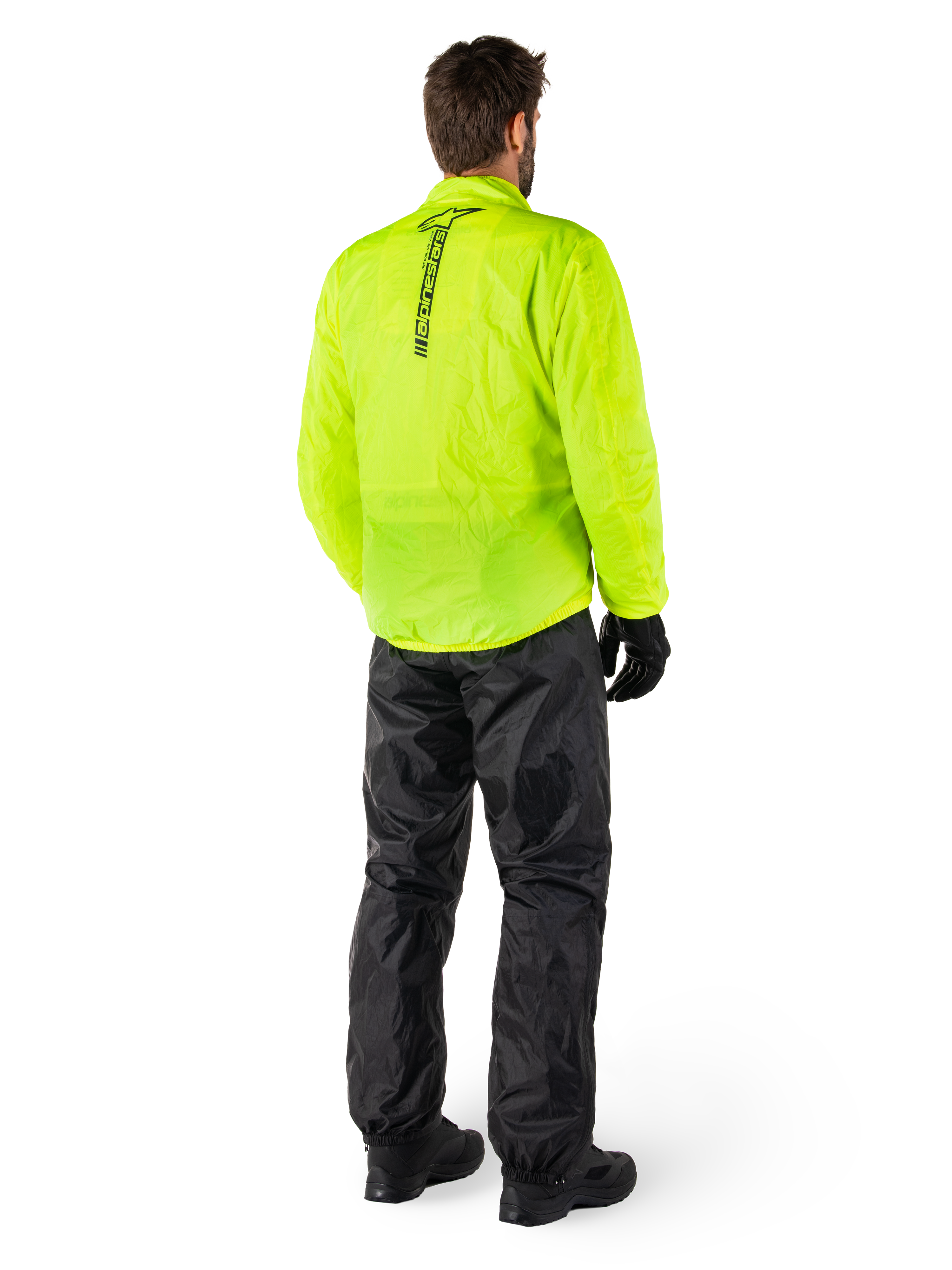 Person von hinten in Alpinestars Hurricane Rain V2 Jacke in Gelb Fluo und Hurricane Rain V2 Hose in Schwarz, vertikales Logo auf dem Jackenrücken, wasserdichte Motorrad-Regenbekleidung mit schwarzen Handschuhen und Stiefeln