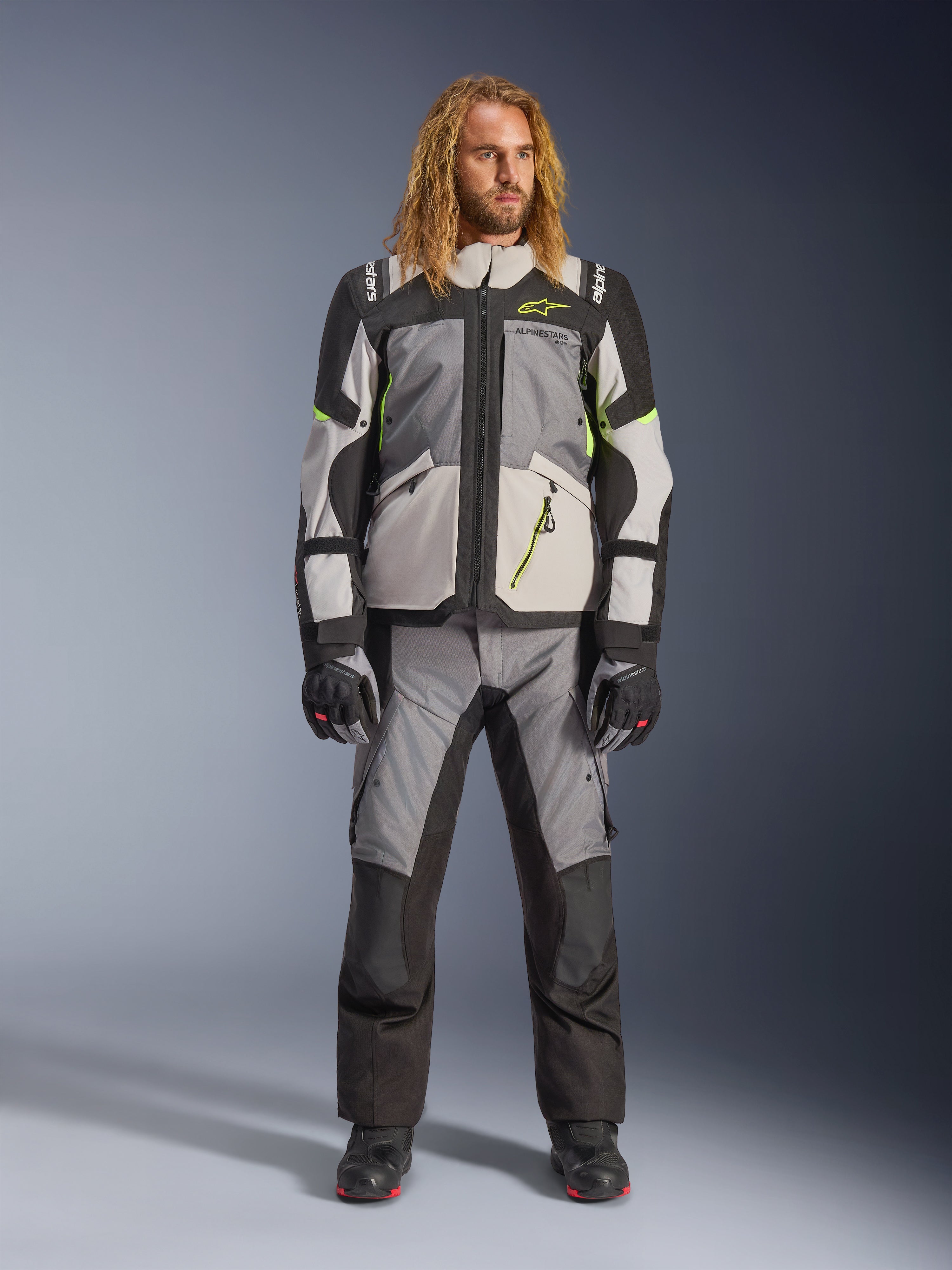 Männliches Model mit langen blonden Haaren in der Andes V4 Drystar® Jacke in Dunkelgrau, Eisgrau, Schwarz und Gelb Fluo, kombiniert mit einer passenden Adventure-Motorradhose und schwarzen Stiefeln, komplette technische Motorradausrüstung für Allwetter-Touren.