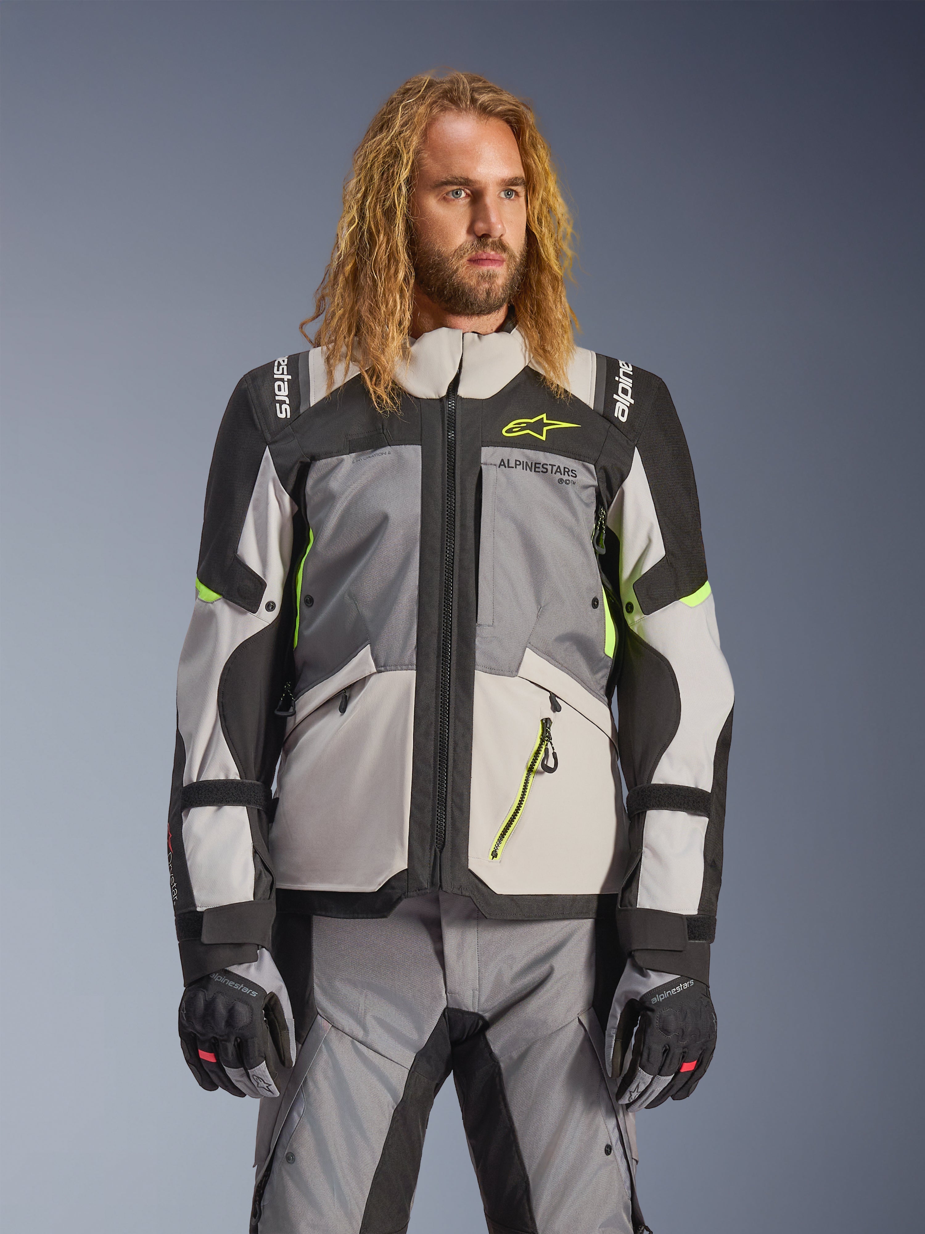 Alpinestars Andes V4 Drystar Jacke, Adventure-Touring-Motorradjacke, Dunkelgrau und Eisgrau mit neongelben Akzenten, getragen von einem Model mit passender Hose und Handschuhen, Multi-Pocket-Textildesign für Fahrten bei jedem Wetter
