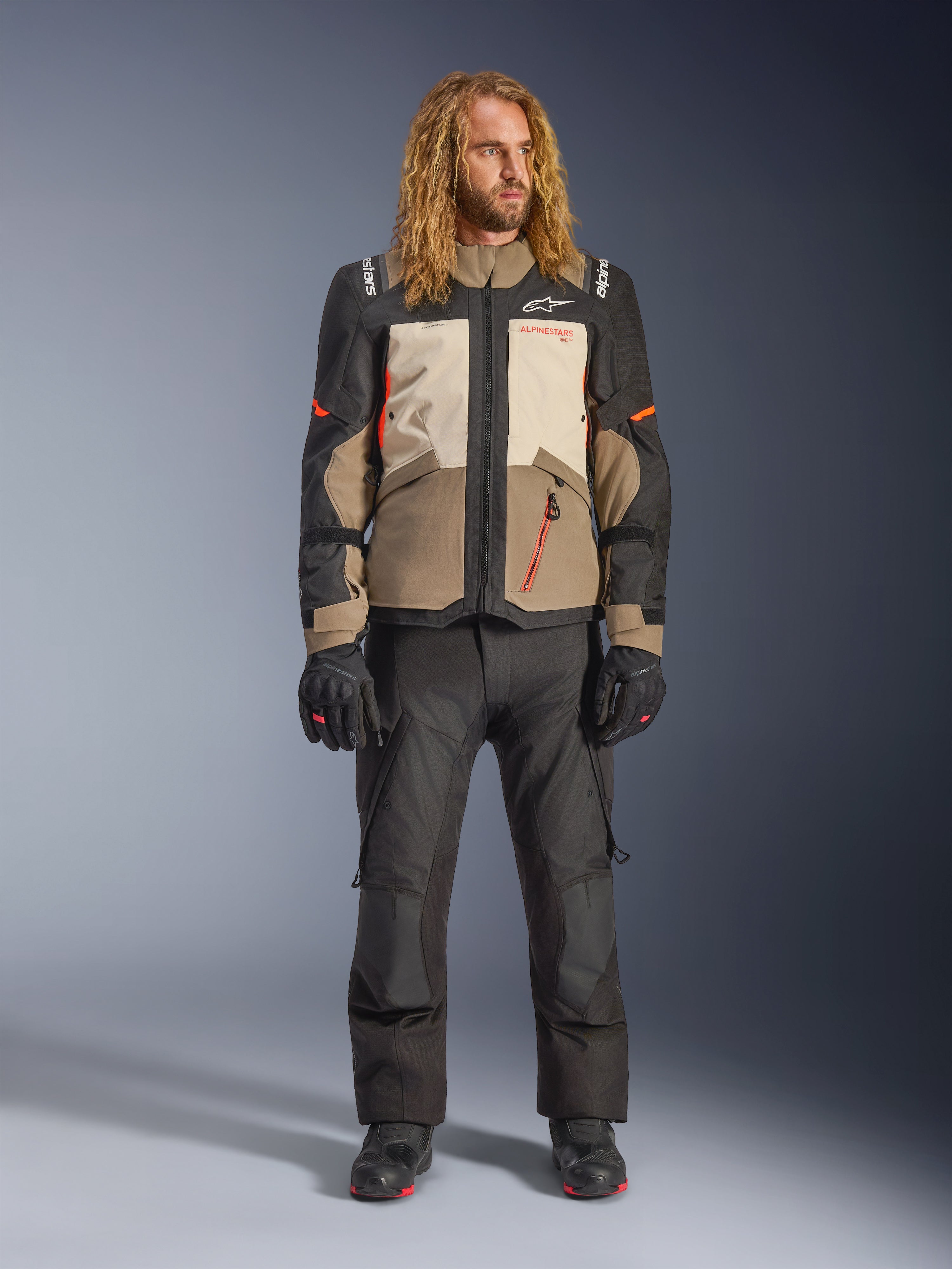 Alpinestars Andes V4 Drystar® Jacket, Walnut Mastic Black, worn with black motorcycle pants and gloves, featuring a multi-panel adventure design with orange accents and Alpinestars branding, technical textile gear for all-weather riding