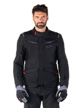 Alpinestars ST-1 Waterproof Jacket, Black motorcycle adventure touring jacket, worn by a male model with matching black pants and grey/black gloves, featuring minimalistic styling, Alpinestars branding on the shoulders, large front cargo pockets with red details, and ergonomic construction for all-weather road touring.