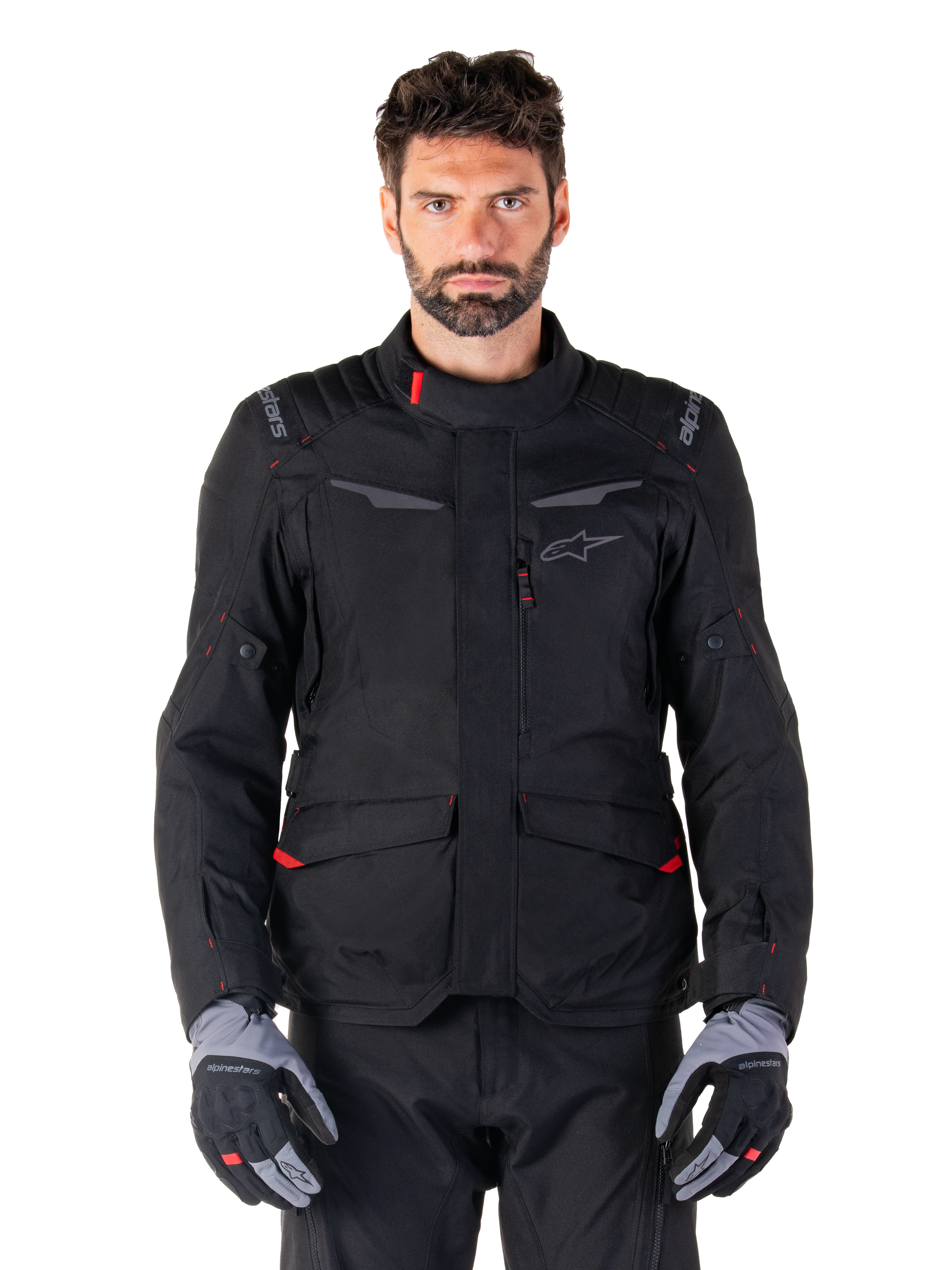 Alpinestars ST-1 Waterproof Jacket, Black motorcycle adventure touring jacket, worn by a male model with matching black pants and grey/black gloves, featuring minimalistic styling, Alpinestars branding on the shoulders, large front cargo pockets with red details, and ergonomic construction for all-weather road touring.