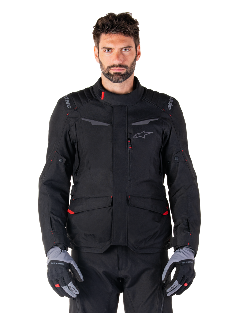 Alpinestars ST-1 Waterproof Jacke, schwarze Motorrad-Adventure-Touring-Jacke, getragen von einem männlichen Model mit passender schwarzer Hose und grau-schwarzen Handschuhen, minimalistisches Design, Alpinestars-Branding an den Schultern, große vordere Cargo-Taschen mit roten Details, ergonomische Konstruktion für Allwetter-Straßentouren