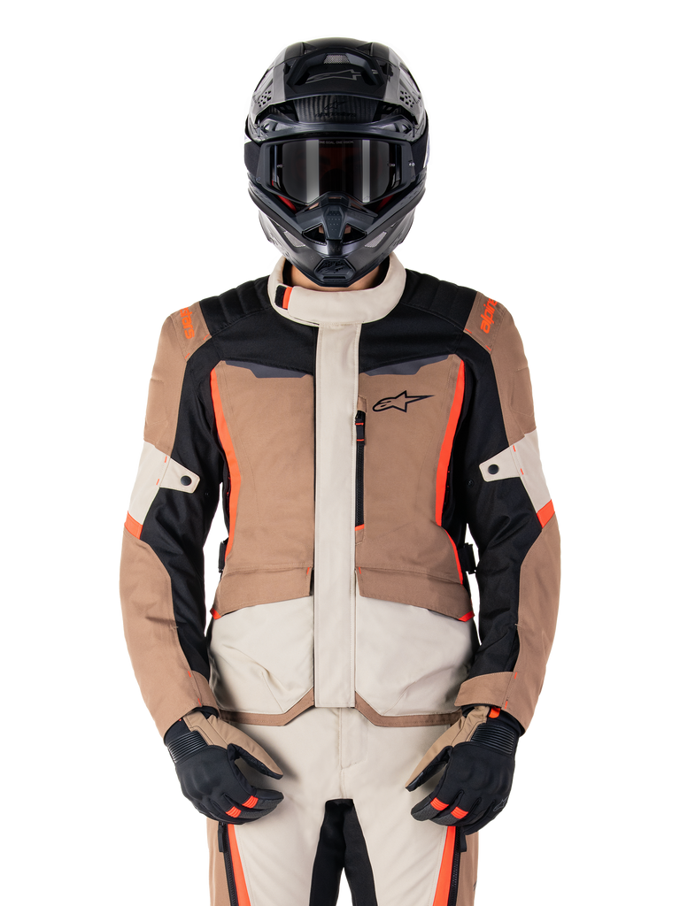 Alpinestars ST-1 Wasserdichte Jacke, Dark Khaki Sand Black Red Fluo, getragen von einem Fahrer mit schwarzem Helm und Handschuhen, Adventure-Touring-Ausrüstung mit beigen und braunen Paneelen und leuchtend roten Fluo-Akzenten, Vorderansicht auf weißem Hintergrund.