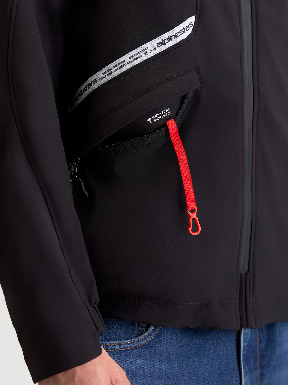 Alpinestars C 5 Drystar Jacke, urbane Herren-Motorradjacke in Schwarz, Nahaufnahme der Keyless-Tasche mit rotem Reißverschlussanhänger und Marken-Brustgrafik