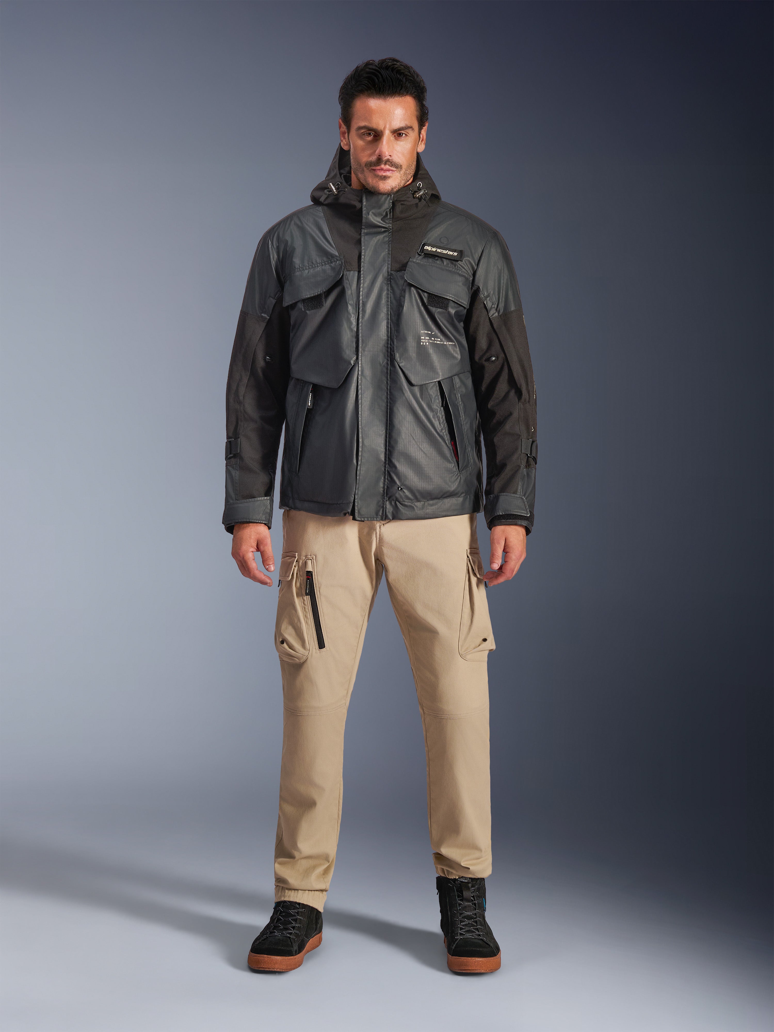 Male model wearing the Alpinestars Mospher Waterproof Jacket in Black, urban contemporary motorcycle gear, tactical utility design with multiple functional pockets and hood, paired with tan cargo pants and black shoes, full-body studio portrait.