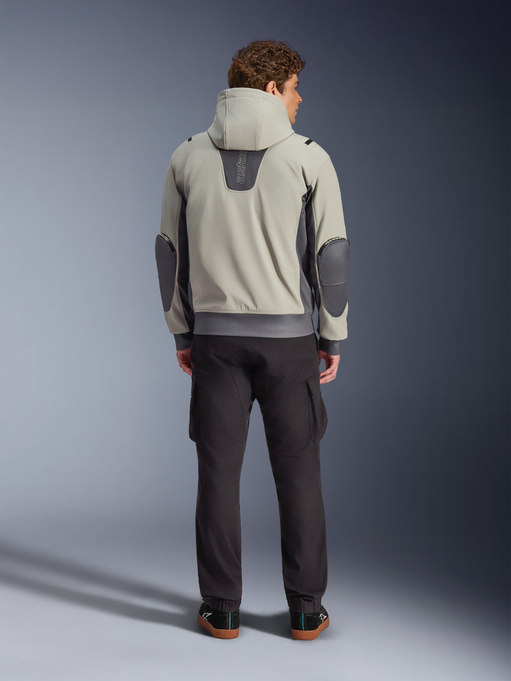 Alpinestars Moblast Wasserdichte Jacke, Iron Gray Dark Sage, Rückansicht eines männlichen Fahrers in der Motorradjacke mit Kapuze, Farbkombination aus Hellgrau und Dark Sage, vertikales „astars“-Logo am oberen Rücken, dunkelgraue Ellbogen-Patches, kombiniert mit dunkler Cargohose für einen modernen urbanen Look.