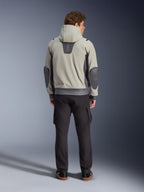 Alpinestars Moblast Wasserdichte Jacke, Iron Gray Dark Sage, Rückansicht eines männlichen Fahrers in der Motorradjacke mit Kapuze, Farbkombination aus Hellgrau und Dark Sage, vertikales „astars“-Logo am oberen Rücken, dunkelgraue Ellbogen-Patches, kombiniert mit dunkler Cargohose für einen modernen urbanen Look.