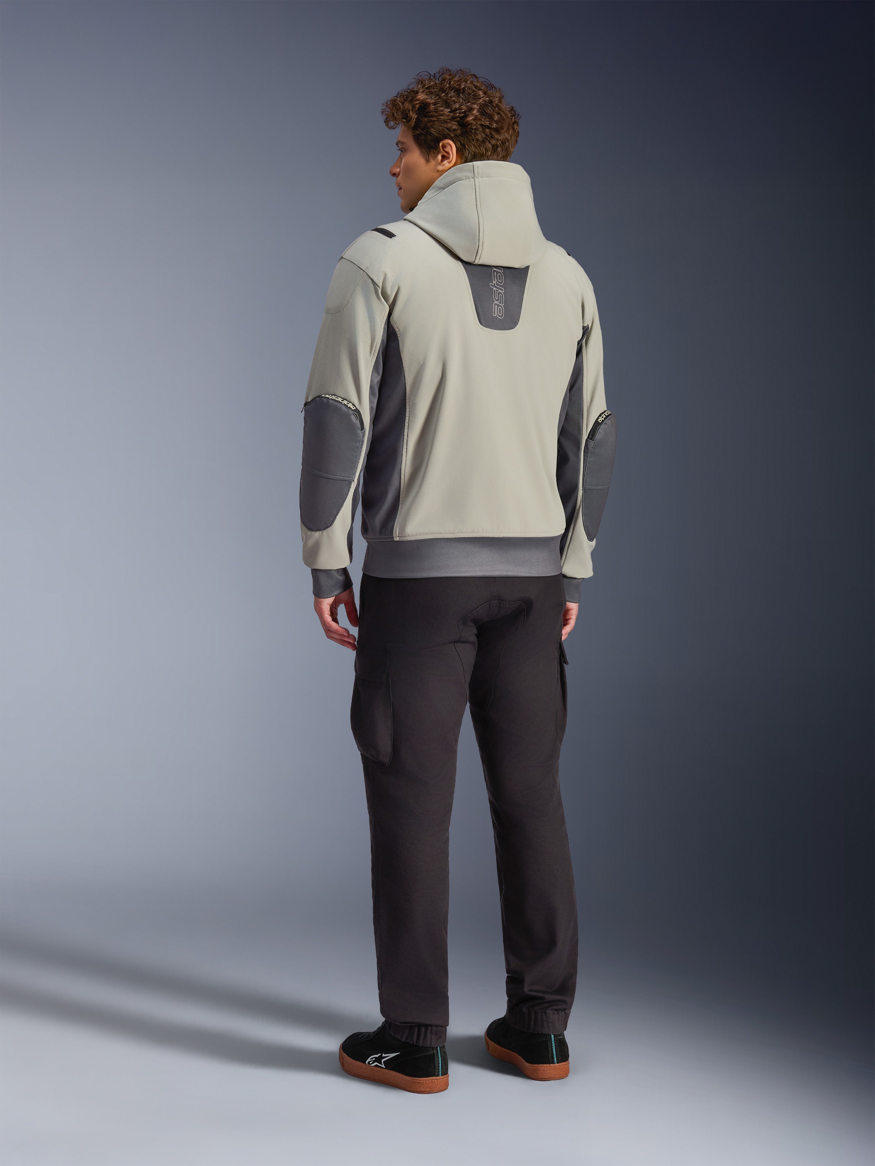 Männliches Model in der Rückansicht, trägt die Alpinestars Moblast Waterproof Jacke in Iron Gray Dark Sage, urbane Motorradjacke mit Kapuze, dunkelgrauen Seiteneinsätzen und verstärkten Ellbogen-Patches, kombiniert mit dunkler Motorradhose und schwarzen Sneakern, moderne minimalistische Ästhetik für den urbanen Pendelverkehr.