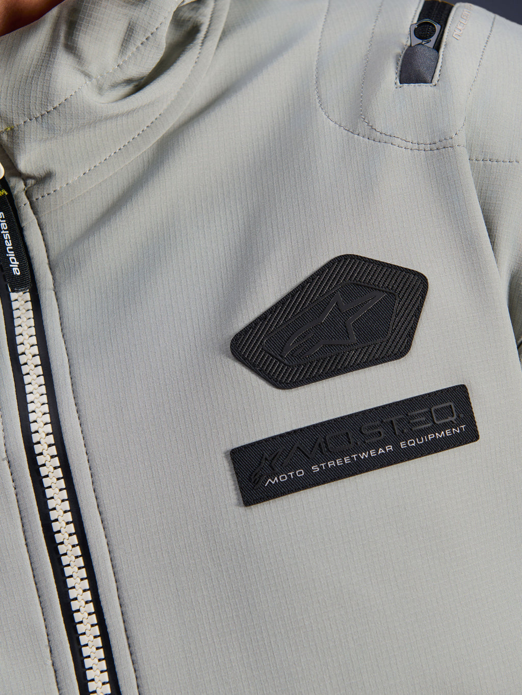 Alpinestars Moblast Wasserdichte Jacke, Iron Gray, Nahaufnahme der Brustaufnäher mit dem Alpinestars Logo und MO.ST.EQ. Moto Streetwear Equipment Branding, technisches Ripstop-Gewebe mit robustem Frontreißverschluss und Detail der Schultertasche.