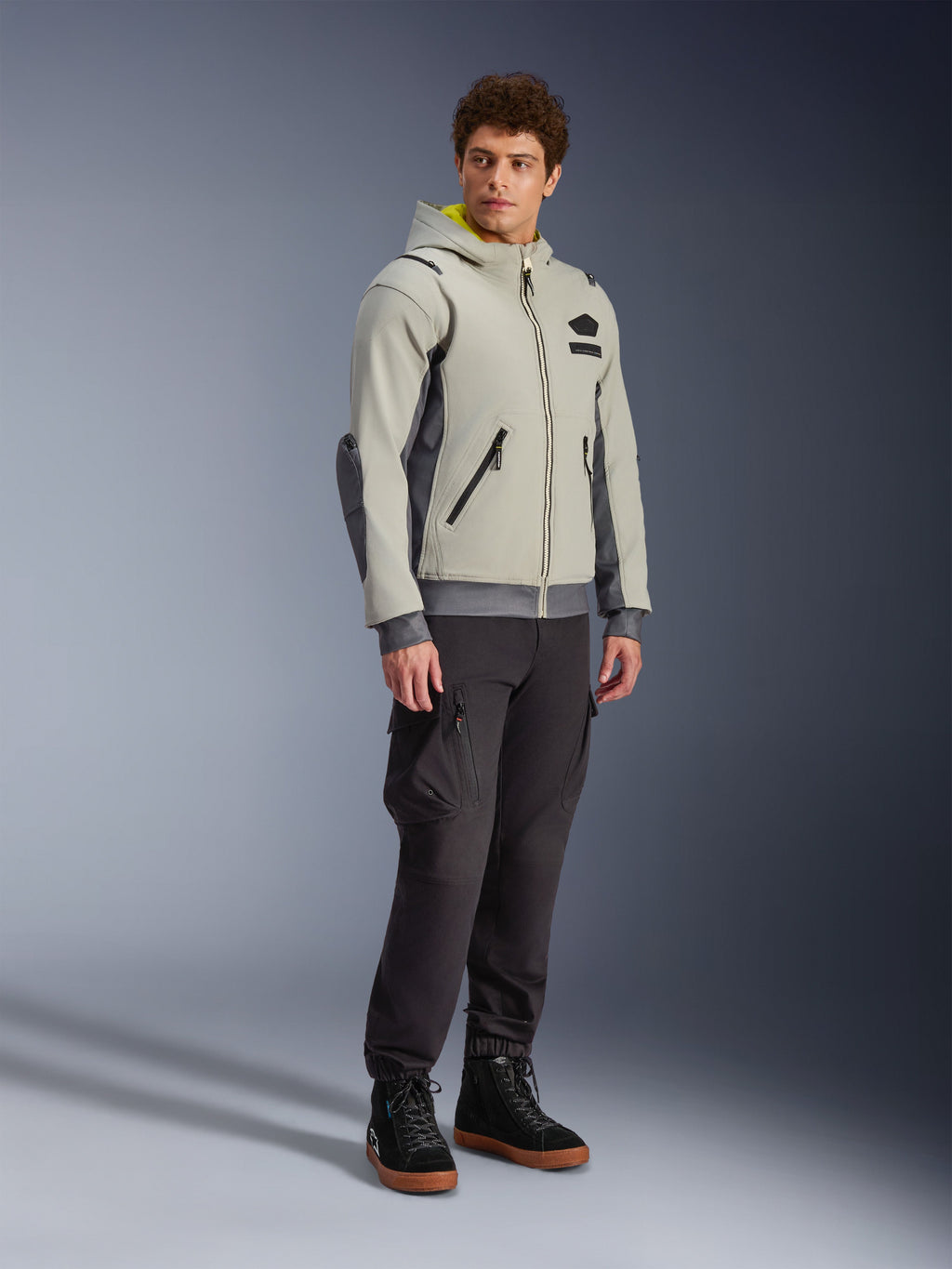 Alpinestars Moblast Wasserdichte Jacke, Iron Gray und Dark Sage, urbane Motorradjacke im Hoodie-Design, getragen von einem Model mit dunkler Motorradhose und schwarzen Stiefeln, minimalistische Cyber-Tech-Ästhetik mit gelber Kapuzeninnenseite und wasserdichten Reißverschlüssen