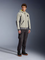 Alpinestars Moblast Wasserdichte Jacke, Iron Gray und Dark Sage, urbane Motorradjacke im Hoodie-Design, getragen von einem Model mit dunkler Motorradhose und schwarzen Stiefeln, minimalistische Cyber-Tech-Ästhetik mit gelber Kapuzeninnenseite und wasserdichten Reißverschlüssen