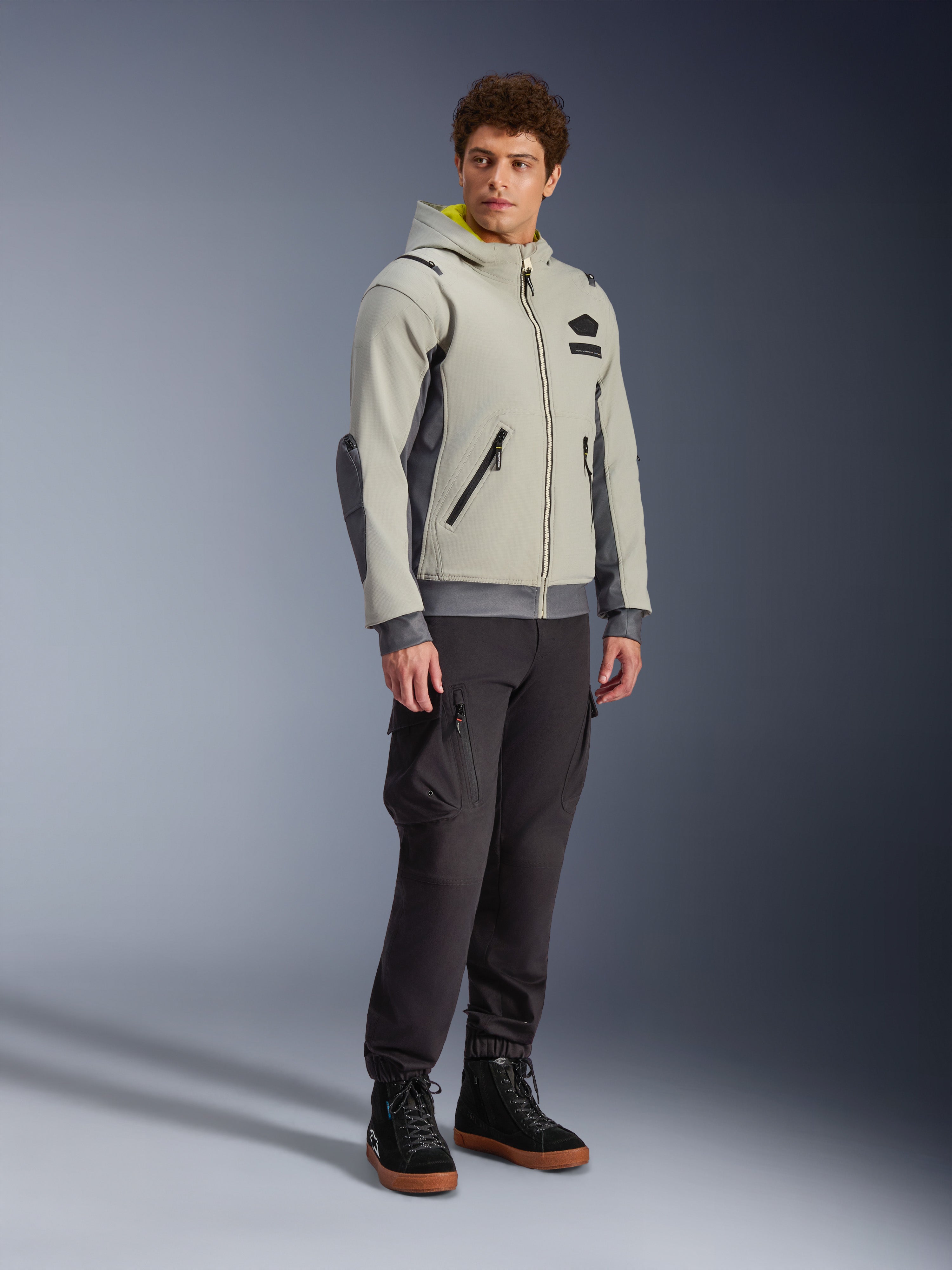 Alpinestars Moblast Wasserdichte Jacke, Iron Gray und Dark Sage, urbane Motorradjacke im Hoodie-Design, getragen von einem Model mit dunkler Motorradhose und schwarzen Stiefeln, minimalistische Cyber-Tech-Ästhetik mit gelber Kapuzeninnenseite und wasserdichten Reißverschlüssen